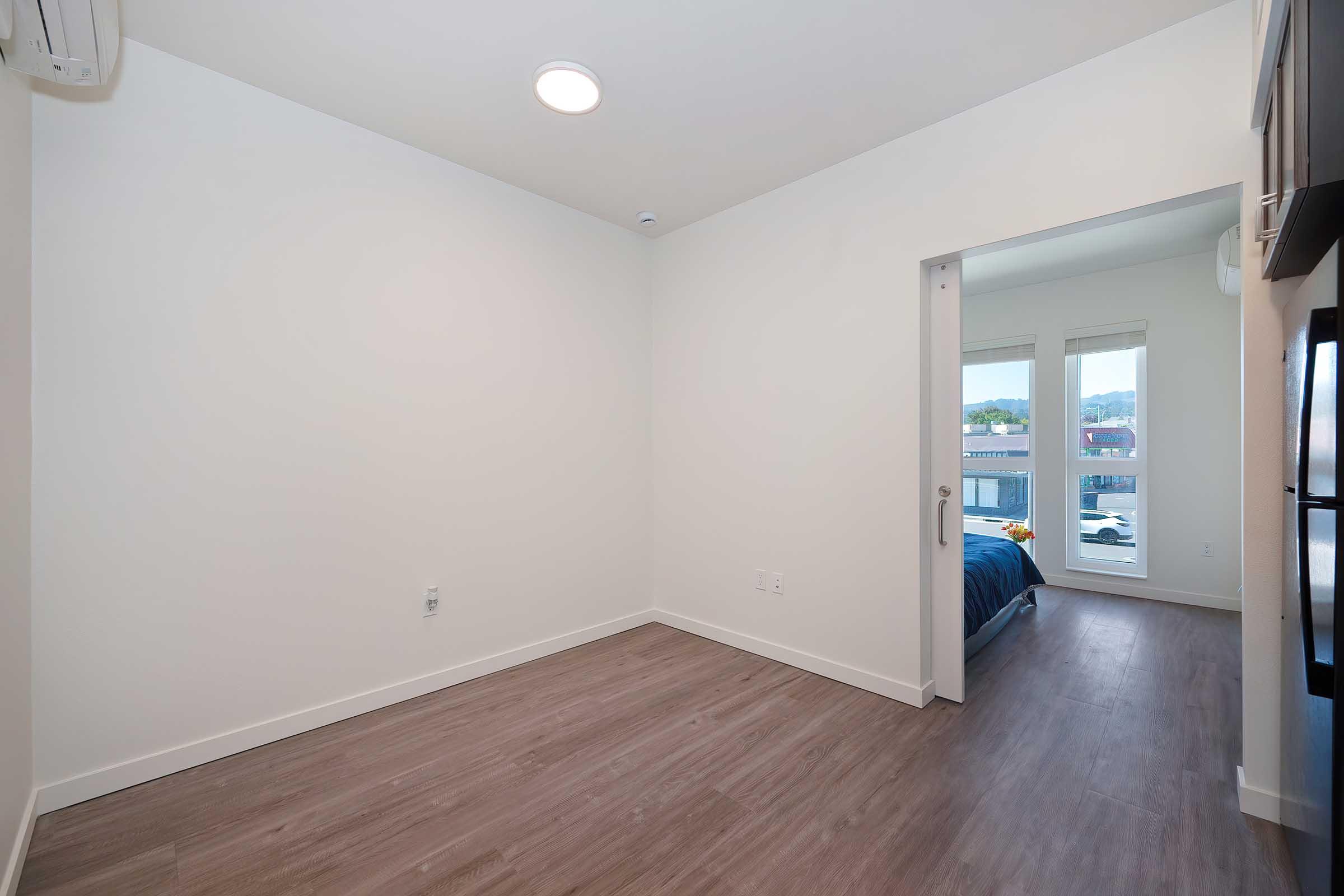 Empty room with light-colored walls and wooden flooring. A door on the right leads to another room with a bed visible through a window. The space has overhead lighting and an air conditioning unit on the wall. Large windows provide natural light, making the room feel bright and open.
