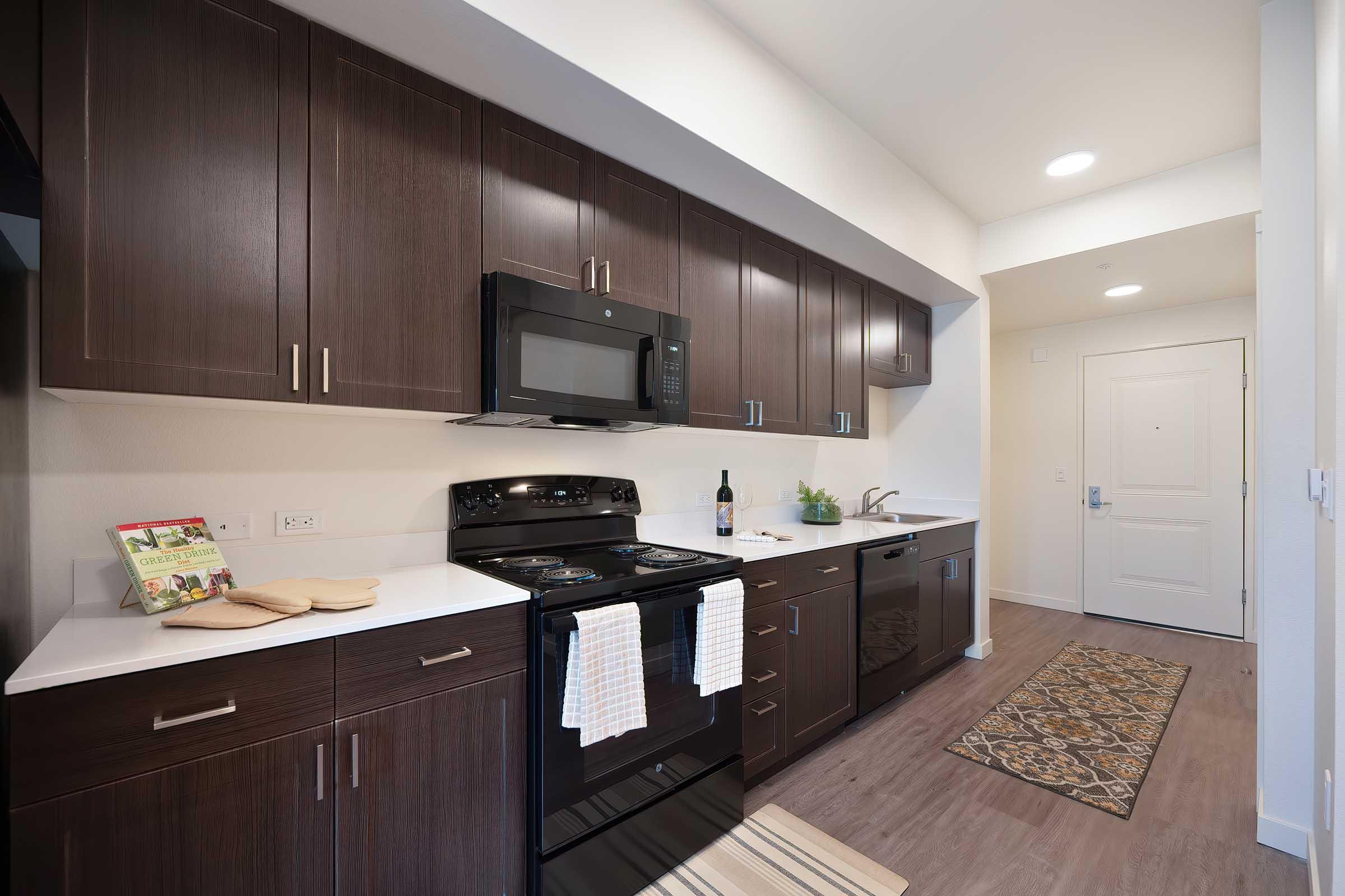 A modern kitchen featuring dark wood cabinets, white countertops, a black stove, an over-the-range microwave, and a dishwasher. A bottle of wine and decorative plant sit on the counter, with a welcome mat near the entrance. The flooring is light wood, and there's a patterned rug by the sink.