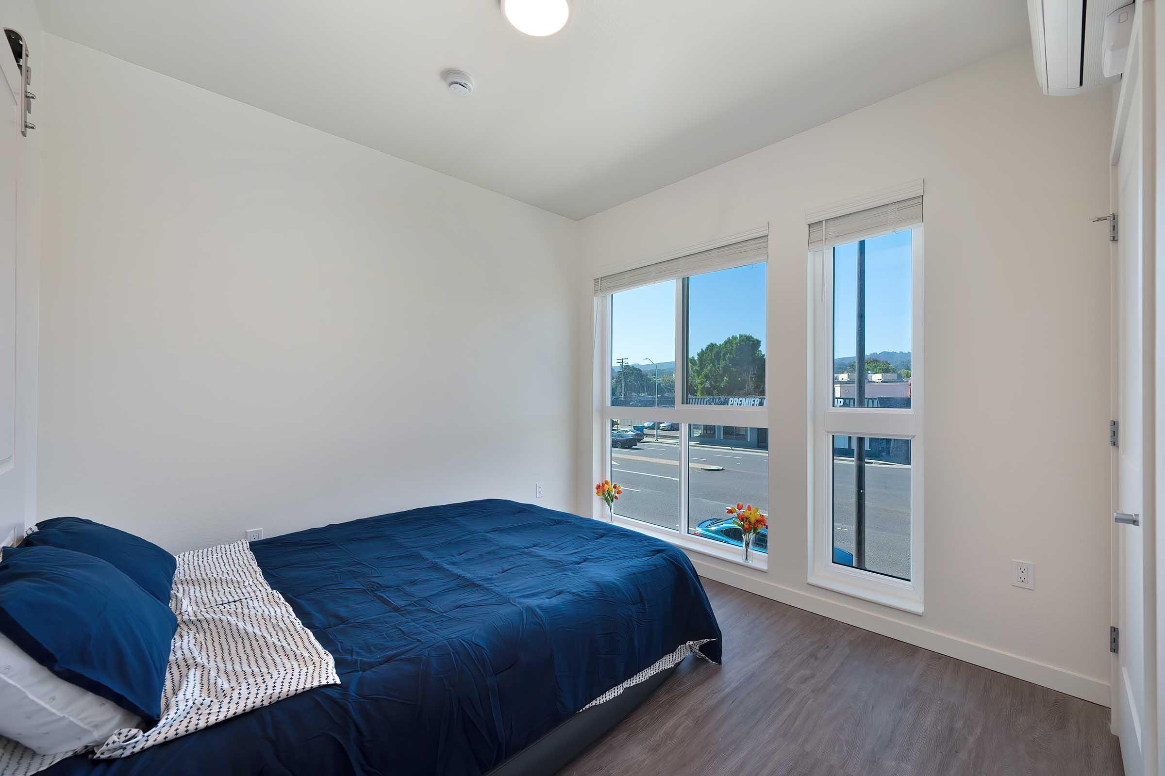 A well-lit bedroom featuring a bed with blue linens, large windows letting in natural light, and a view of the outdoors. The room has a minimalistic aesthetic with light walls and a sleek floor, complemented by potted flowers on the window sill.