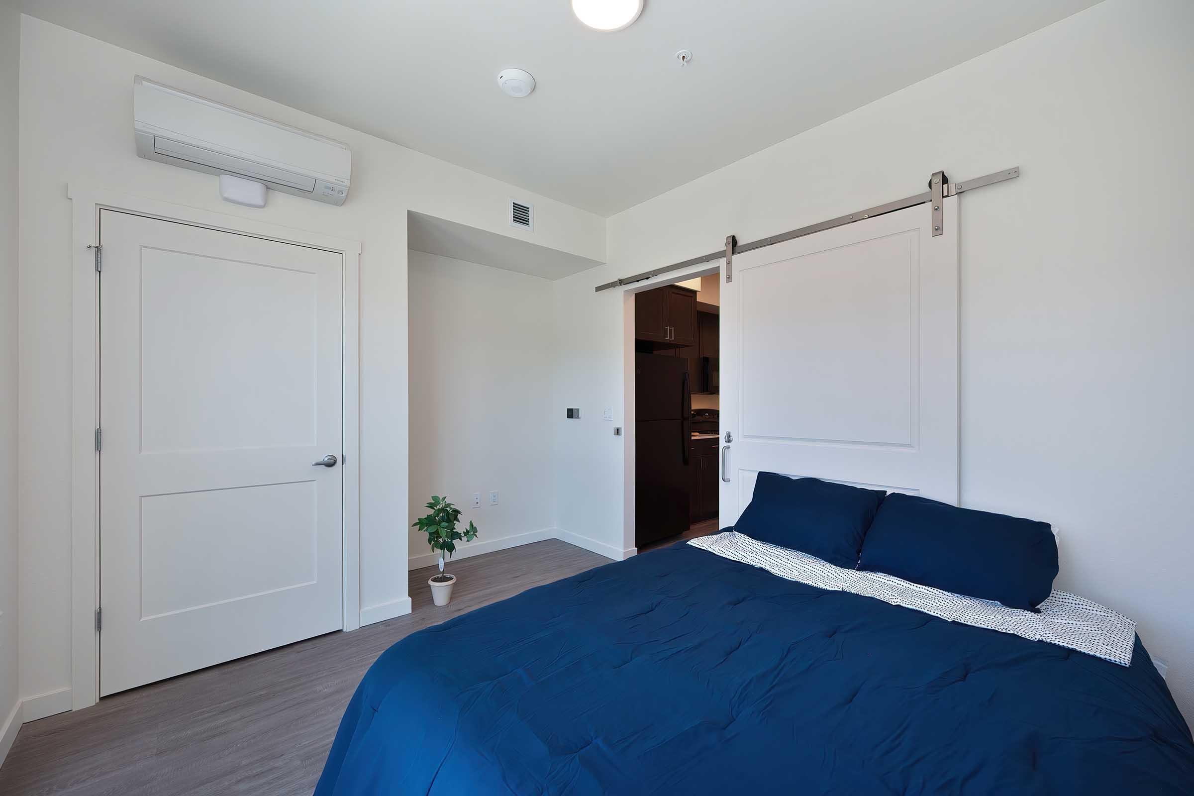 A modern bedroom featuring a blue bedspread on a neatly made bed, a small potted plant on the floor, light-colored walls, and sleek sliding barn doors. An air conditioning unit is mounted on the wall, and a doorway leads to a small kitchenette area. The room has a clean and minimalistic design.