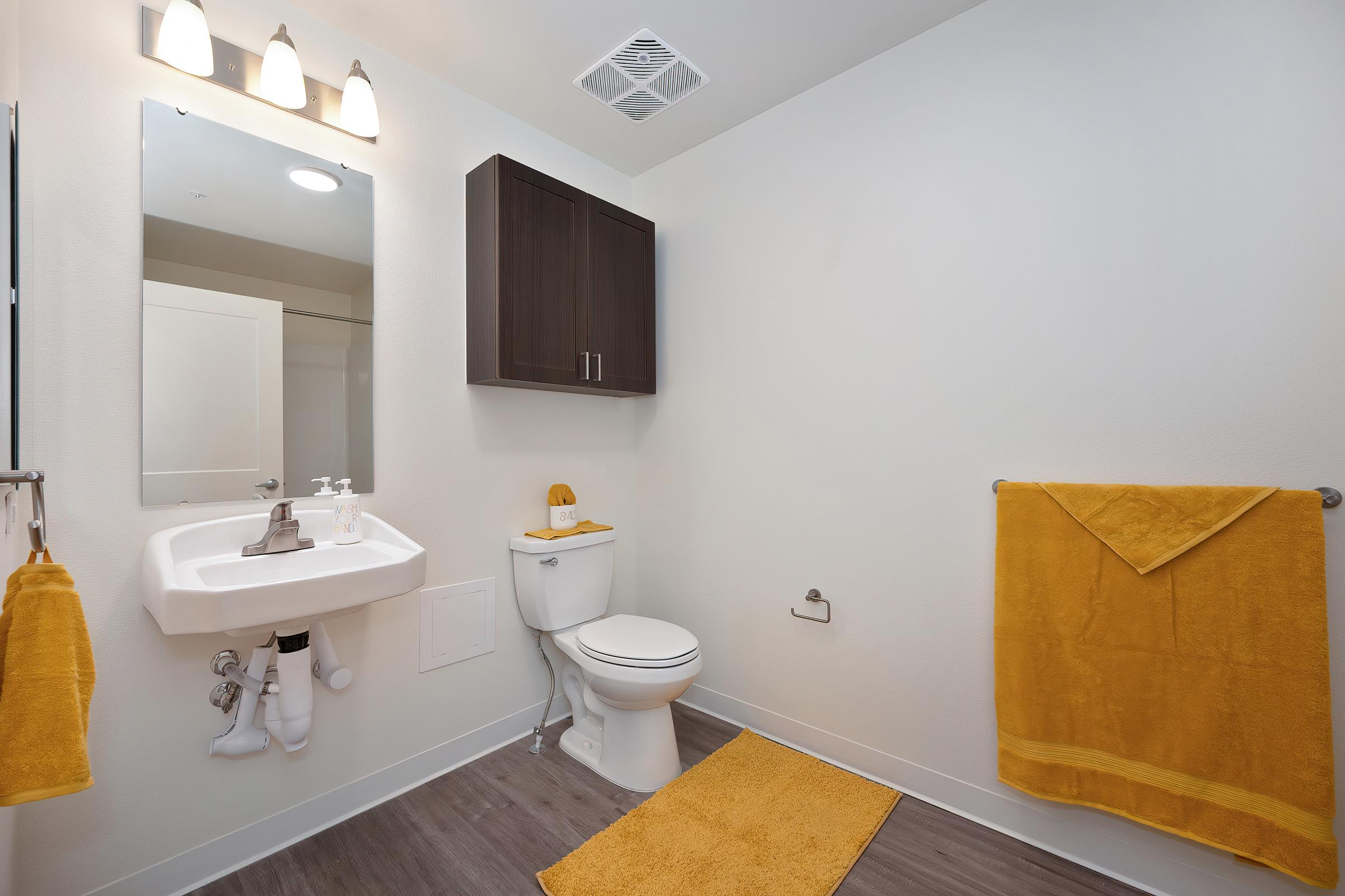 A modern bathroom featuring a white toilet, a small sink with a mirror above it, and dark wood cabinets. Yellow towels and a bath mat add a pop of color. The walls are painted light, and the flooring is a stylish dark wood laminate. Ceiling lights illuminate the space.