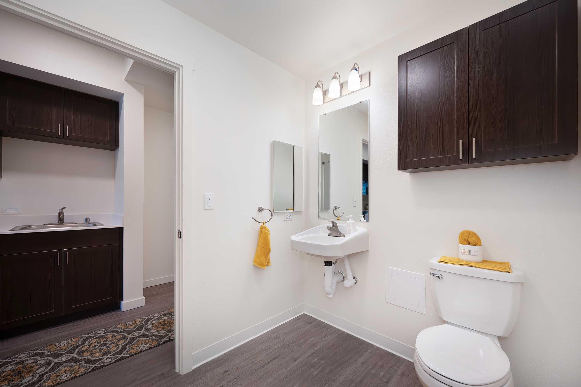 A modern bathroom featuring a white toilet, a sleek sink with a mirror above, and cabinetry with dark wood finishes. A yellow towel and decorative item add a pop of color, while the light walls and flooring create a bright atmosphere. A small kitchenette is visible in the background.