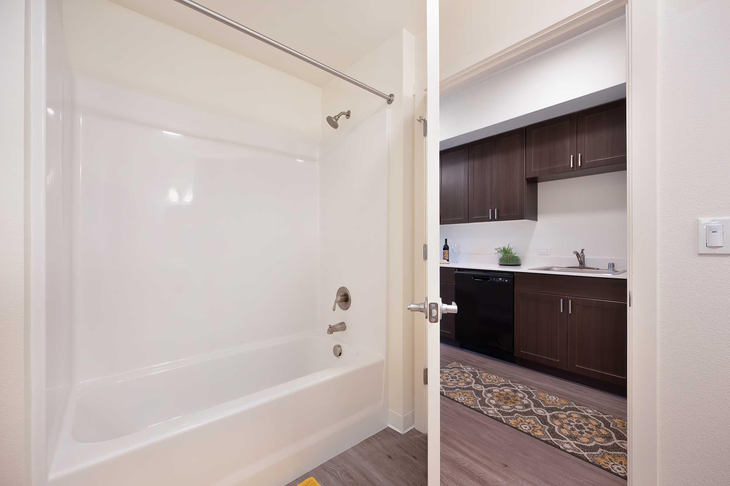A clean, modern bathroom featuring a white bathtub and shower, partially open door leading to a kitchen area with dark wood cabinetry and a small plant on the counter. The kitchen is equipped with a black dishwasher and a decorative rug on the floor.