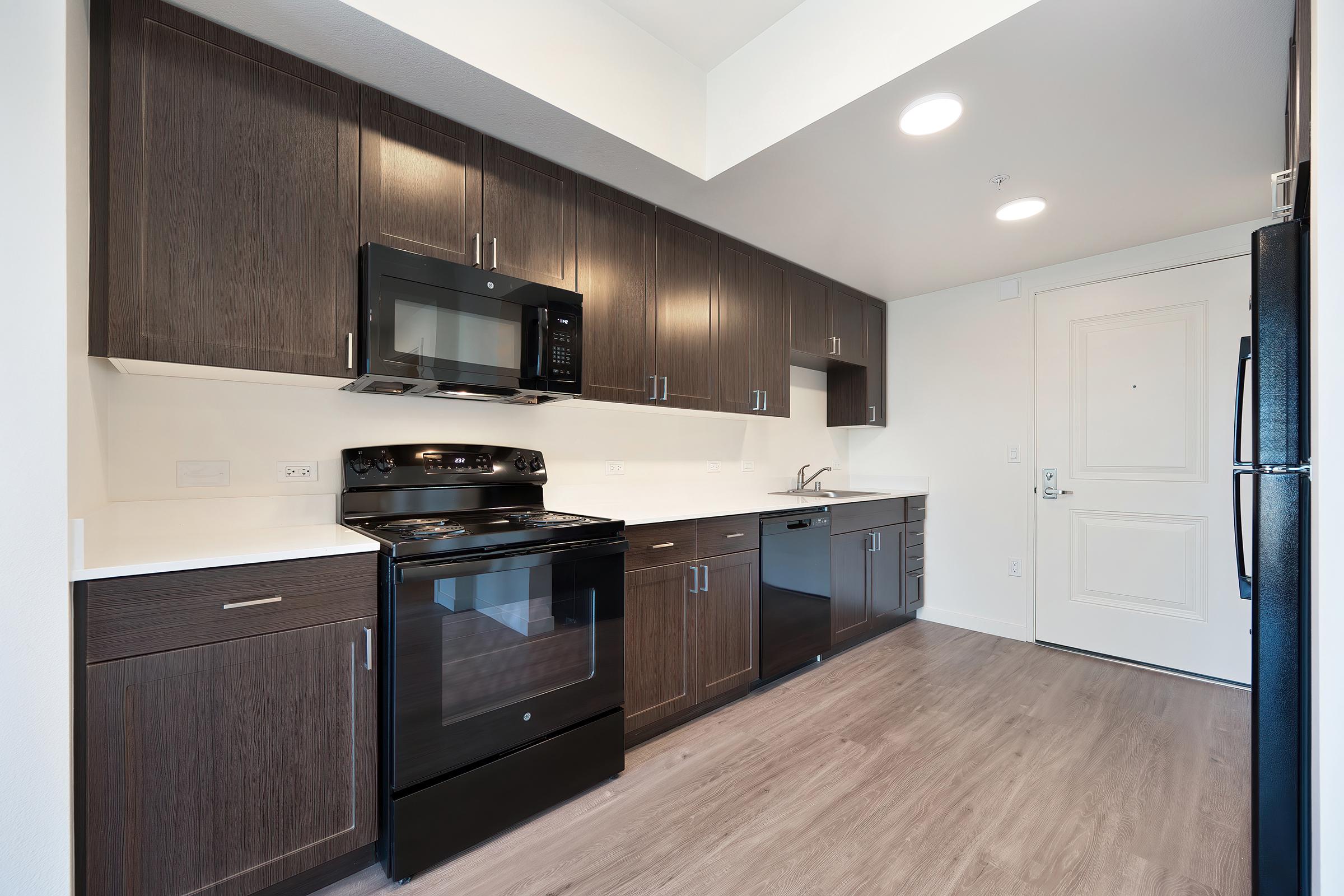 Modern kitchen featuring dark wood cabinetry, stainless steel appliances including a microwave, oven, and dishwasher, a white countertop, and a door leading to another room. The floor is light wood, with overhead recessed lighting that brightens the space.