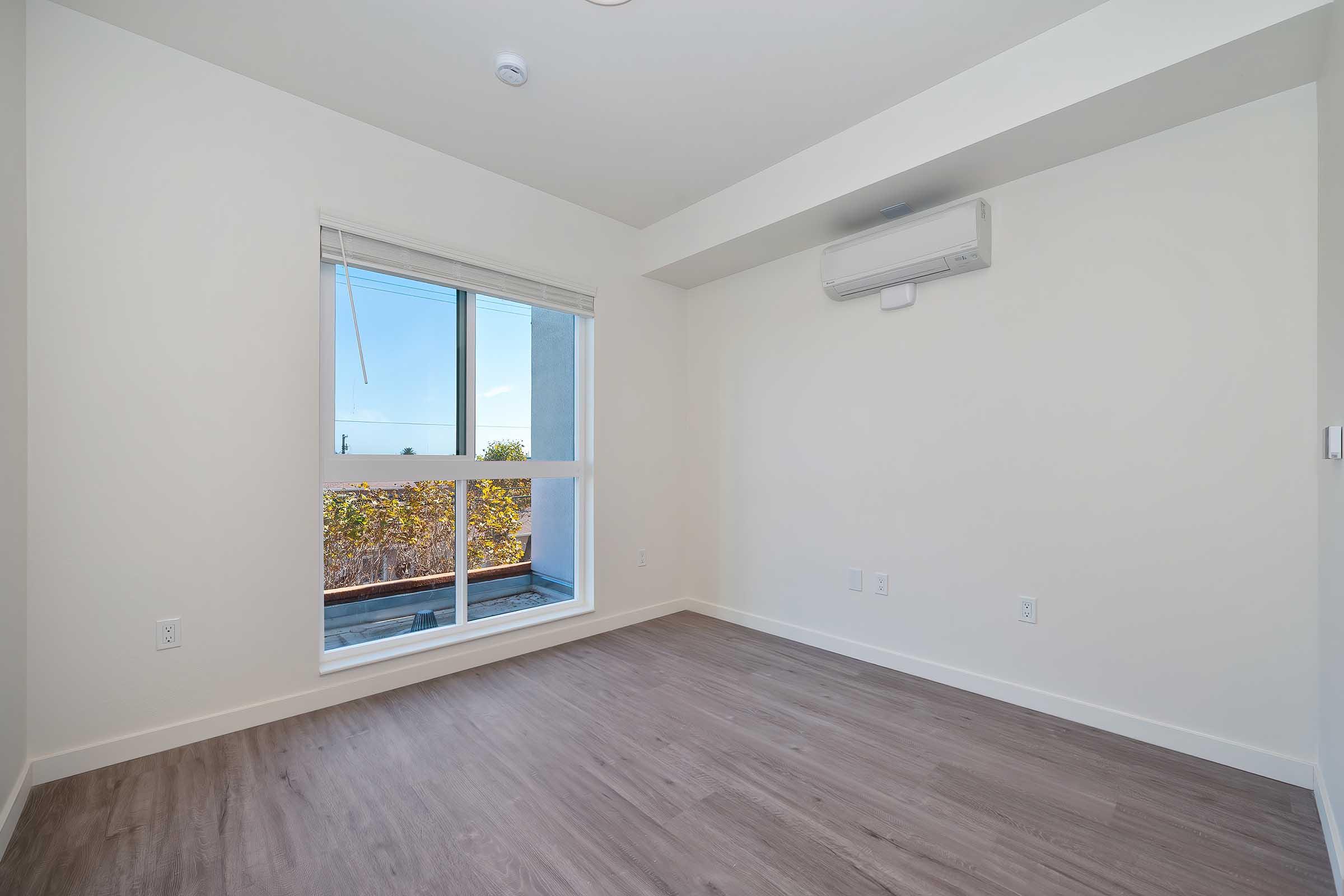A spacious, empty room featuring light-colored walls and hardwood flooring. A large window lets in natural light, offering a view of greenery outside. An air conditioning unit is mounted on the wall, contributing to the room's modern design.