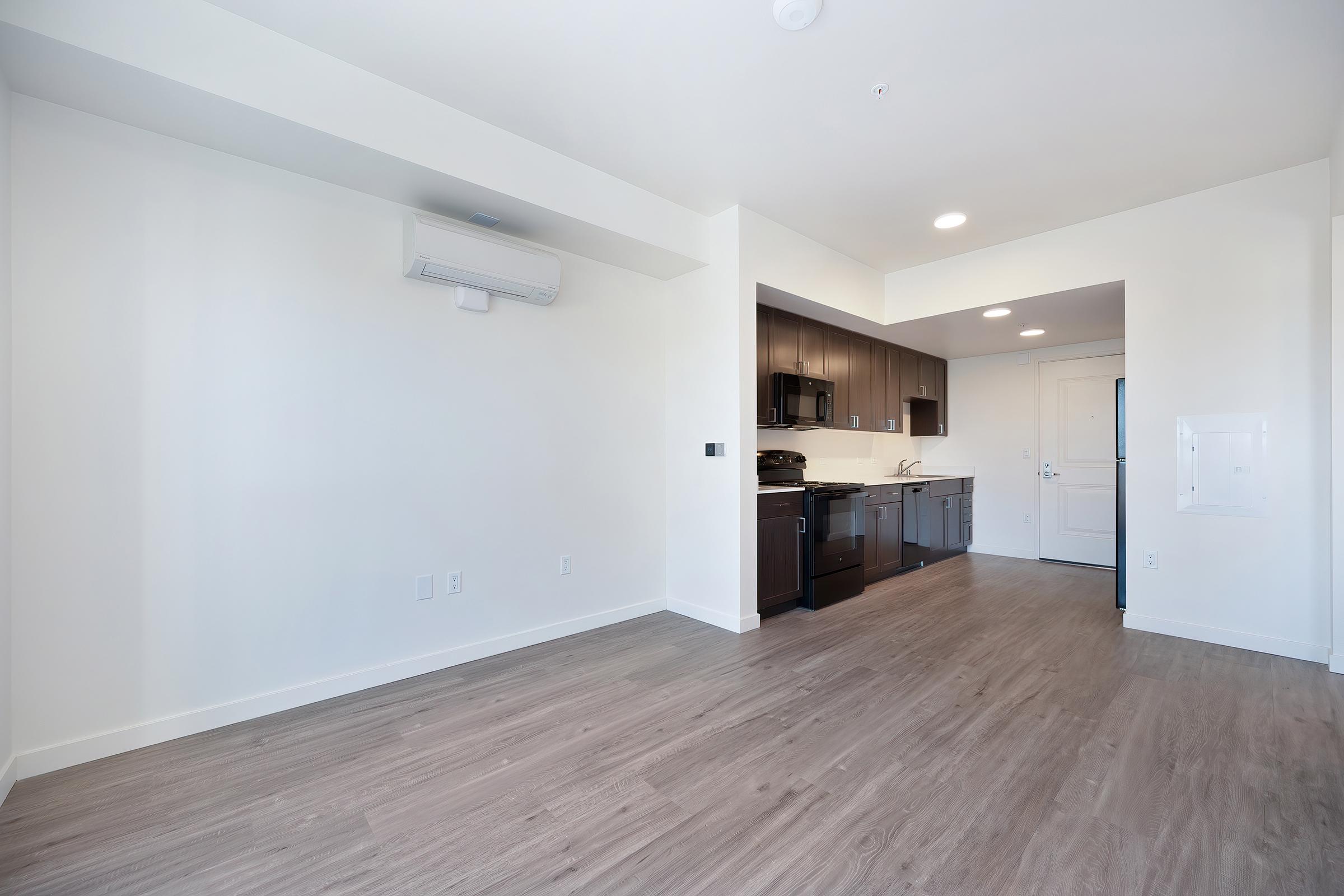 A bright, modern studio apartment featuring an open layout with light wood flooring. The kitchen is visible in the background, equipped with dark cabinetry and stainless steel appliances. The living area has white walls and a wall-mounted air conditioner, creating a spacious and inviting atmosphere.