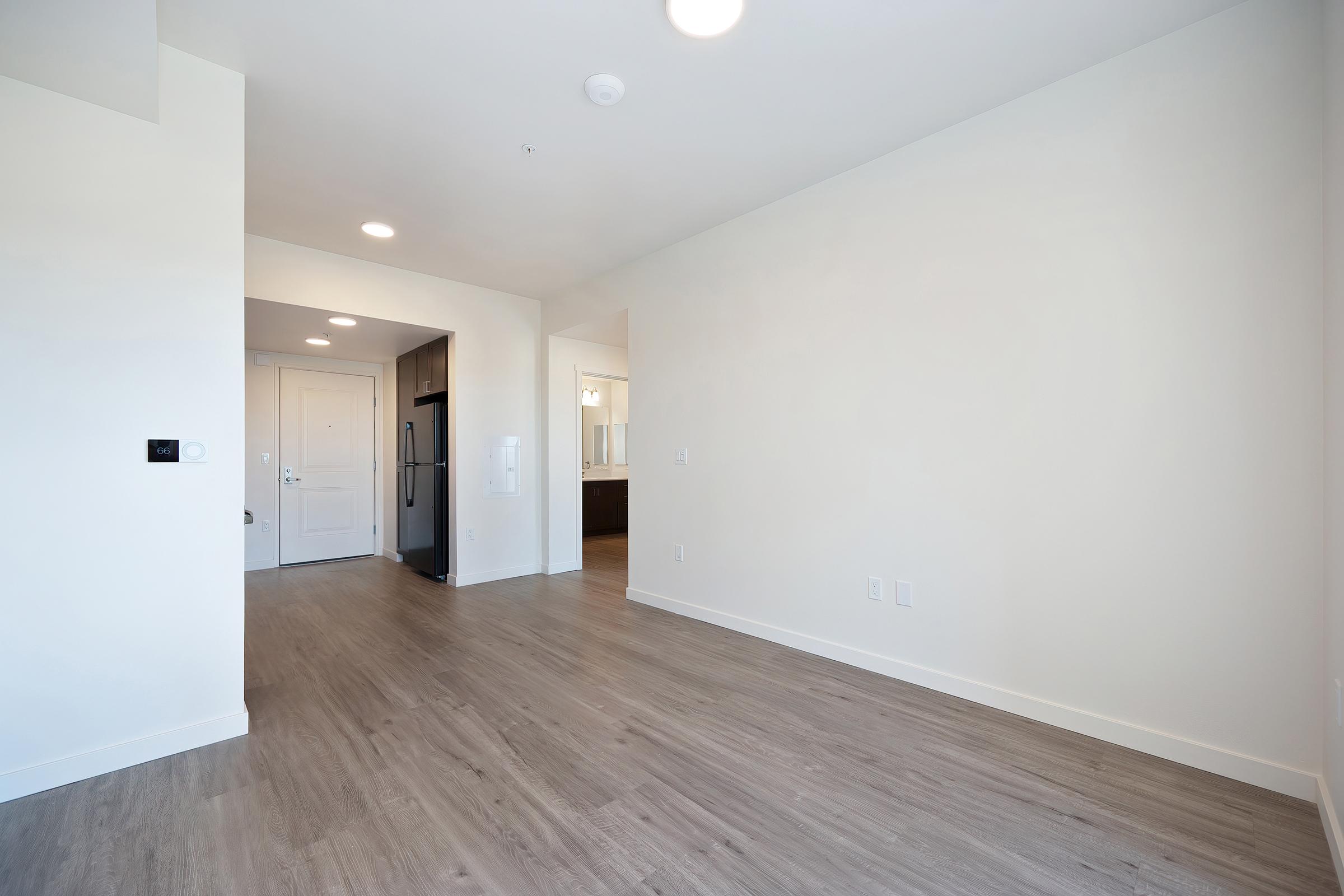 A spacious, unfurnished room with light-colored walls and wood laminate flooring. The entryway features a white door and framed doorway leading to a kitchen area with dark cabinetry and appliances visible. Soft lighting from recessed fixtures illuminates the space, creating a bright and airy feel.