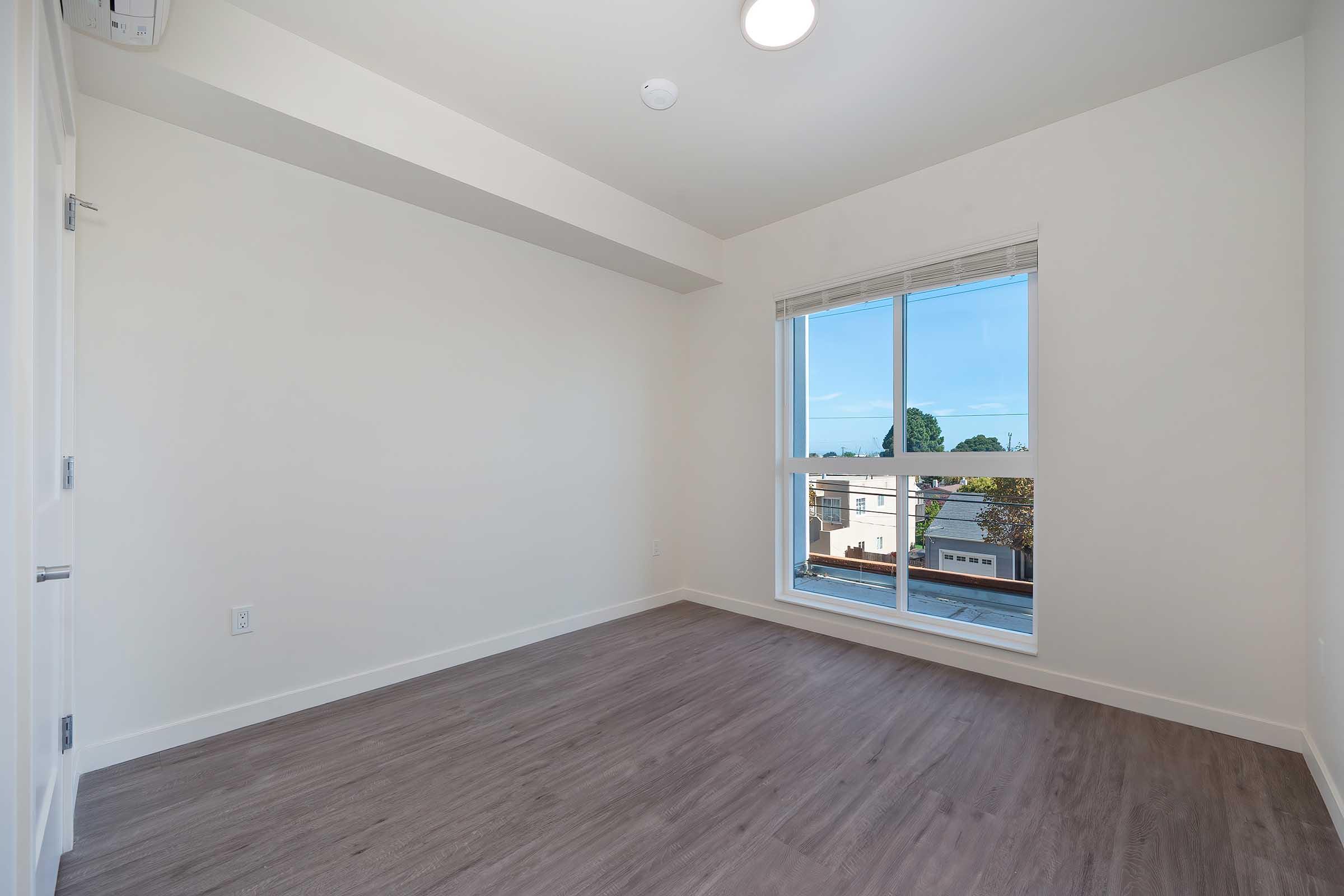 A spacious, empty room with light-colored walls and a large window letting in natural light. The floor features light brown laminate. The view outside includes rooftops and trees. The ceiling has a circular light fixture, adding to the bright ambiance.