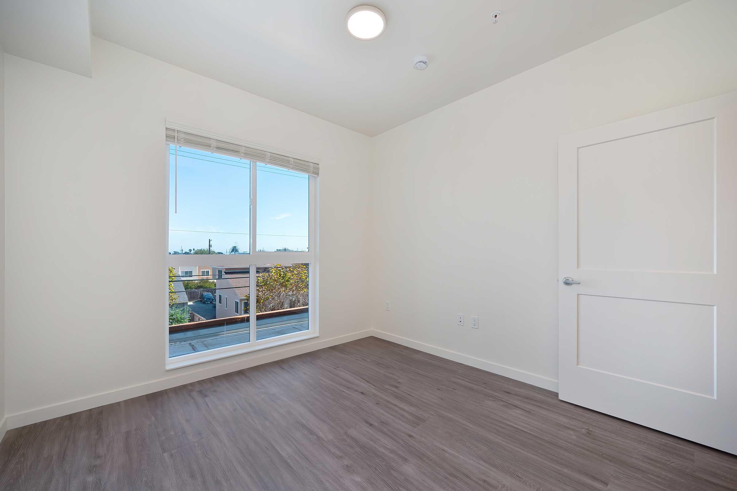 Empty room with light-colored walls and a large window allowing natural light. The floor has a wood-like finish, and there is a white door on the right. The view outside the window shows a clear sky. The space is uncluttered, presenting a modern and minimalist aesthetic.