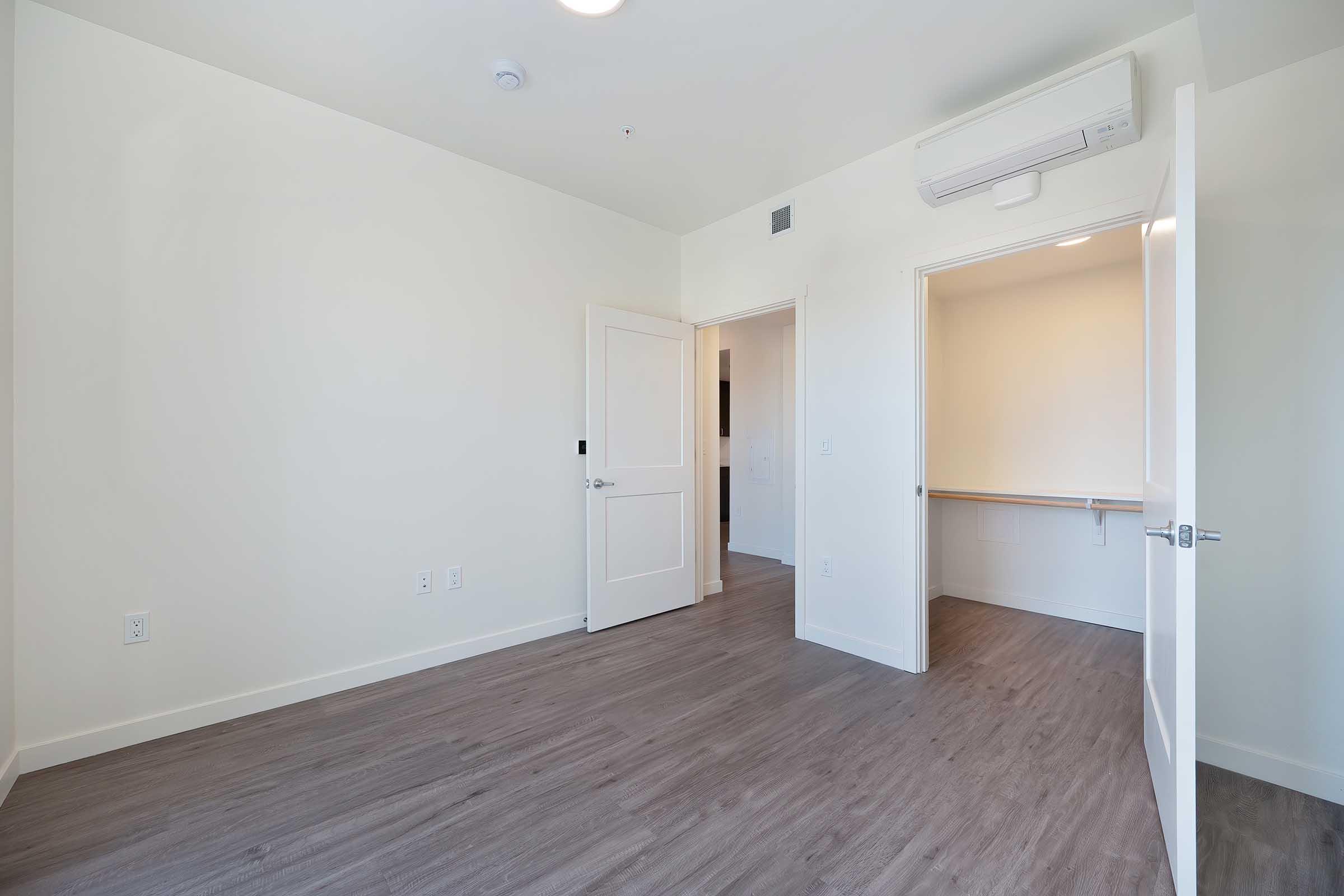A spacious, well-lit room with light-colored walls, a wooden floor, and a ceiling-mounted air conditioning unit. Two open doors lead to adjoining areas, and there is a closet space visible on the right. The overall look is modern and minimalistic, suitable for a bedroom or personal office.