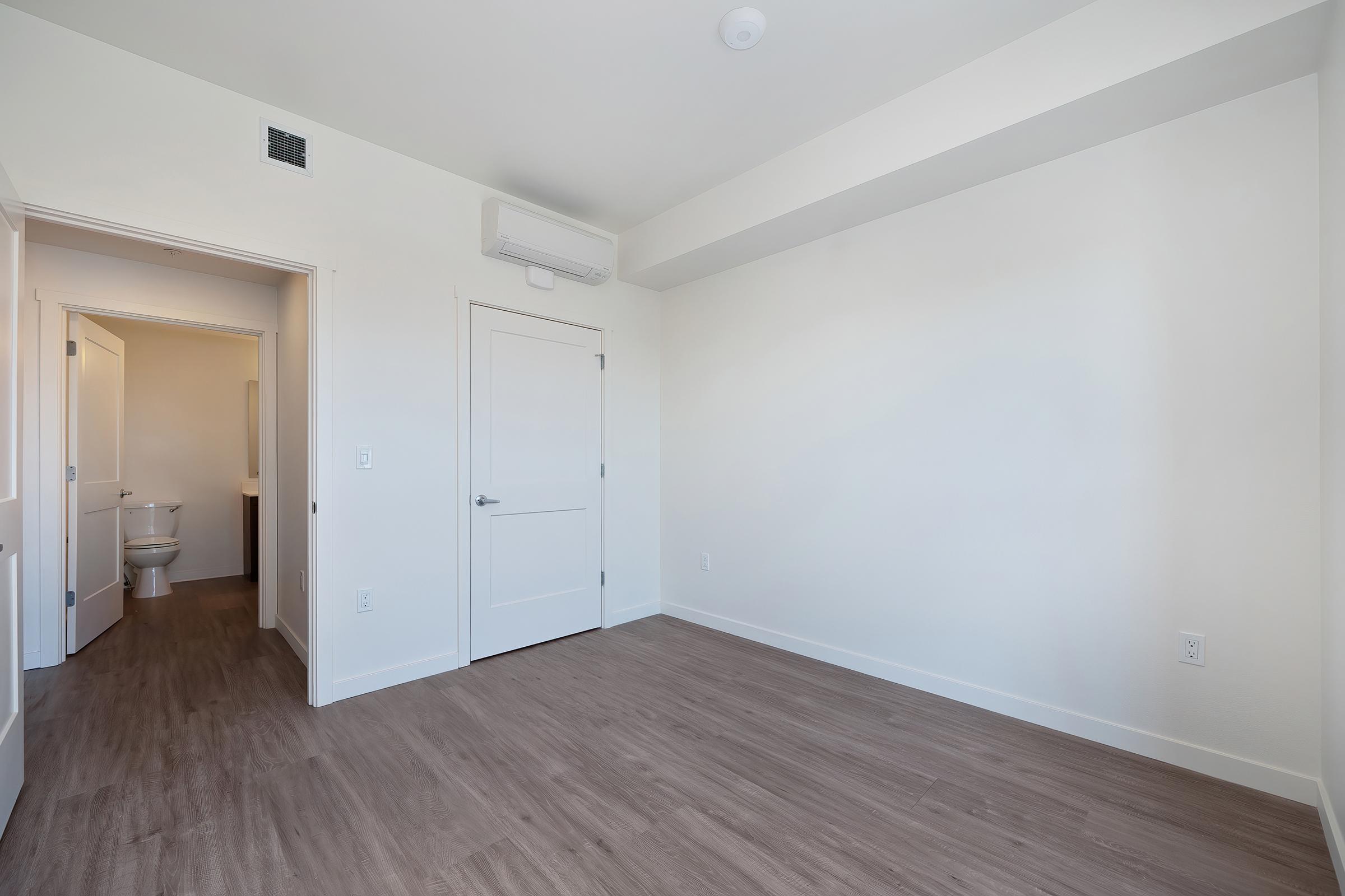 A spacious, empty room with light-colored walls and wooden flooring. There are two doorways leading to adjacent rooms, including one that appears to lead to a bathroom. A wall-mounted air conditioning unit is installed near the ceiling. Natural light brightens the room through an unobtrusive window.