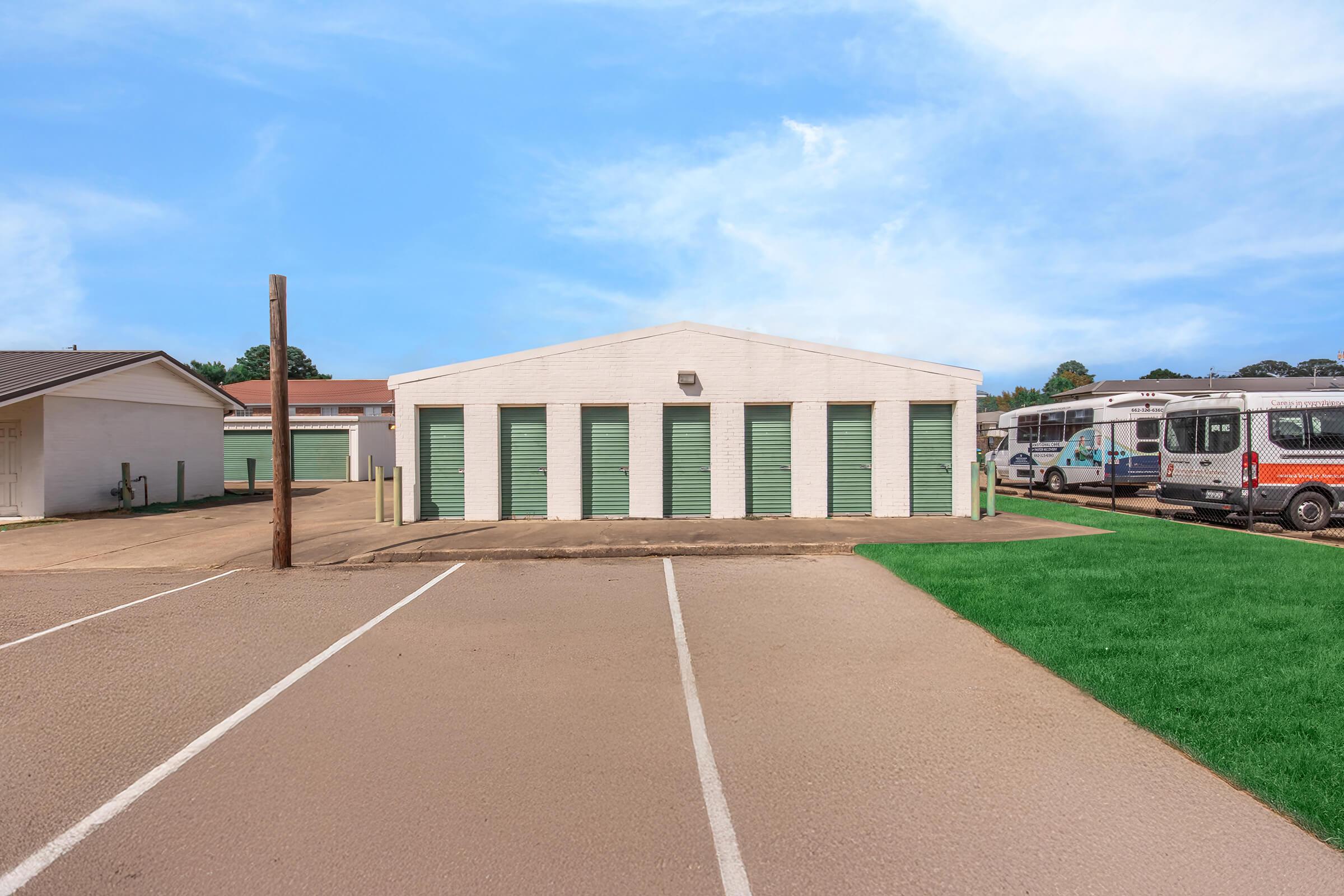 A storage facility featuring a row of storage units with green roll-up doors, set against a clear blue sky. In the foreground, there is an empty parking lot, and to the side, there are vehicles parked, including a bus and a van. The surrounding area includes green grass and a few buildings in the background.