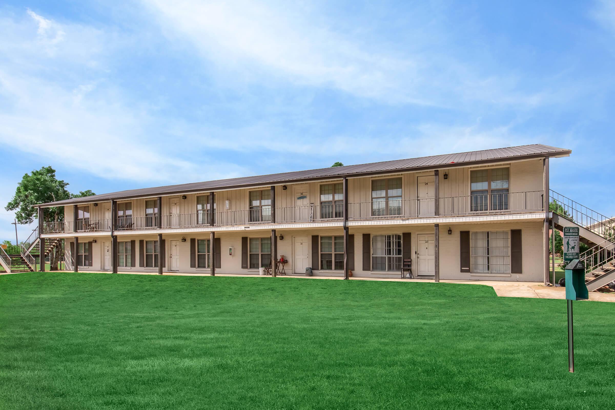 A two-story motel or apartment building with multiple units. Each unit features a small porch with railings, and the exterior is painted in a light color. The building is set against a clear blue sky, and there's a well-maintained green lawn in front.