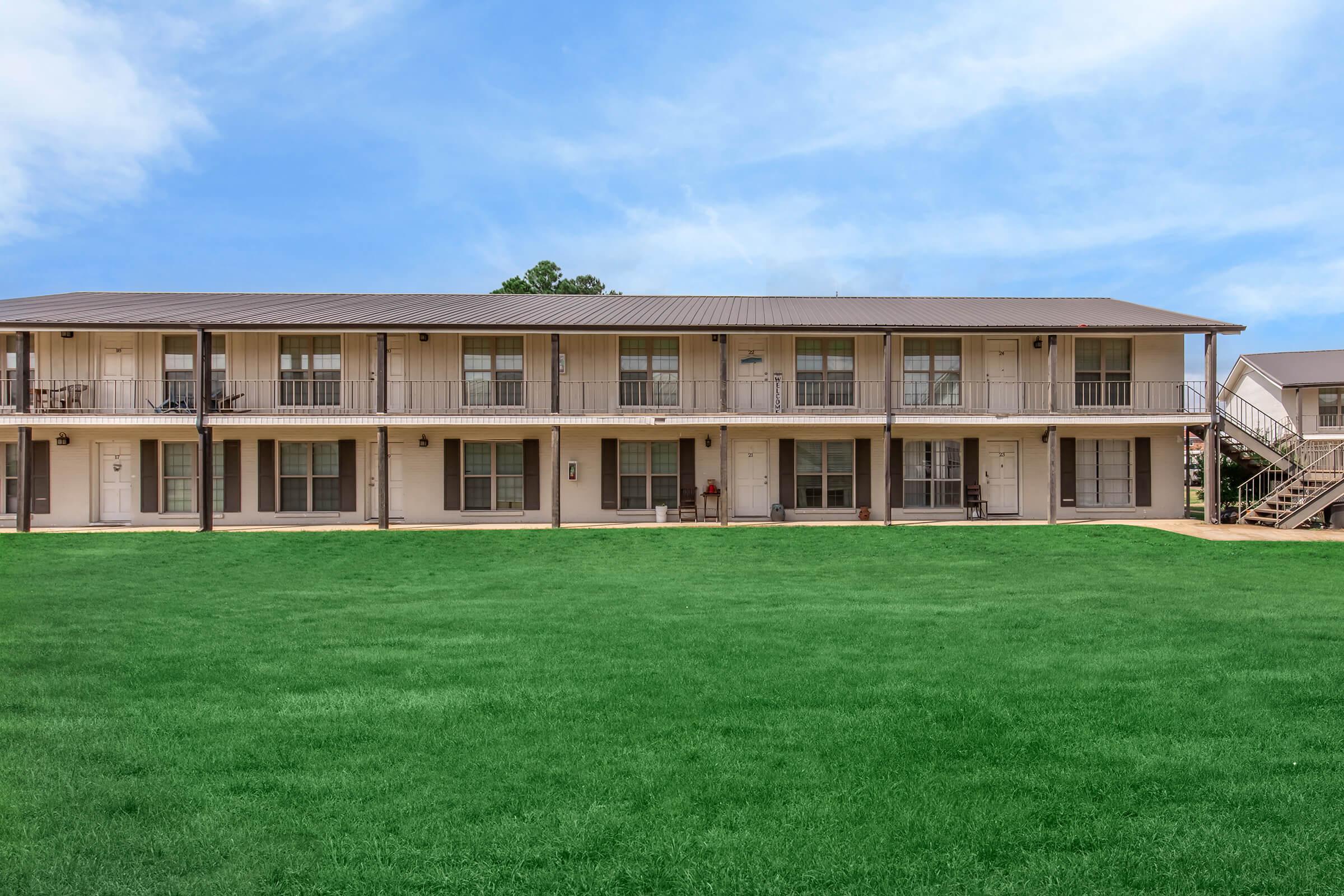 Two-story apartment building with multiple units, featuring a light-colored exterior and balconies. The building is surrounded by a lush green lawn, and the sky above is clear and blue with a few clouds. Paths lead to the entrance of the units.