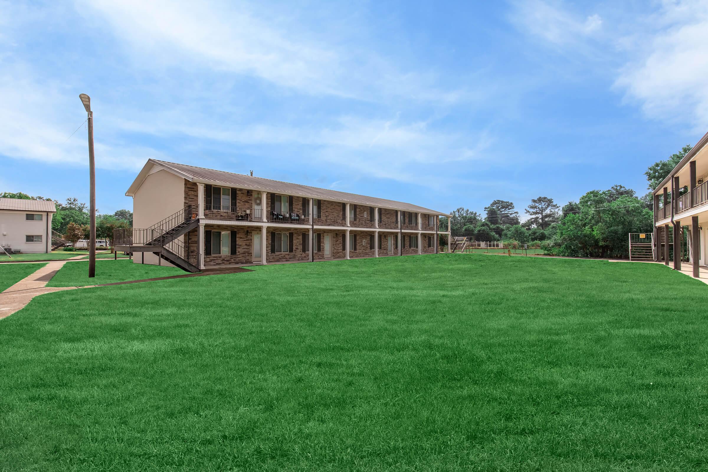 A wide view of a two-story building with stone and beige exterior, featuring several balconies. The building faces a spacious green lawn under a clear blue sky. A pathway runs alongside the building, and a few trees are visible in the background, creating a serene environment.