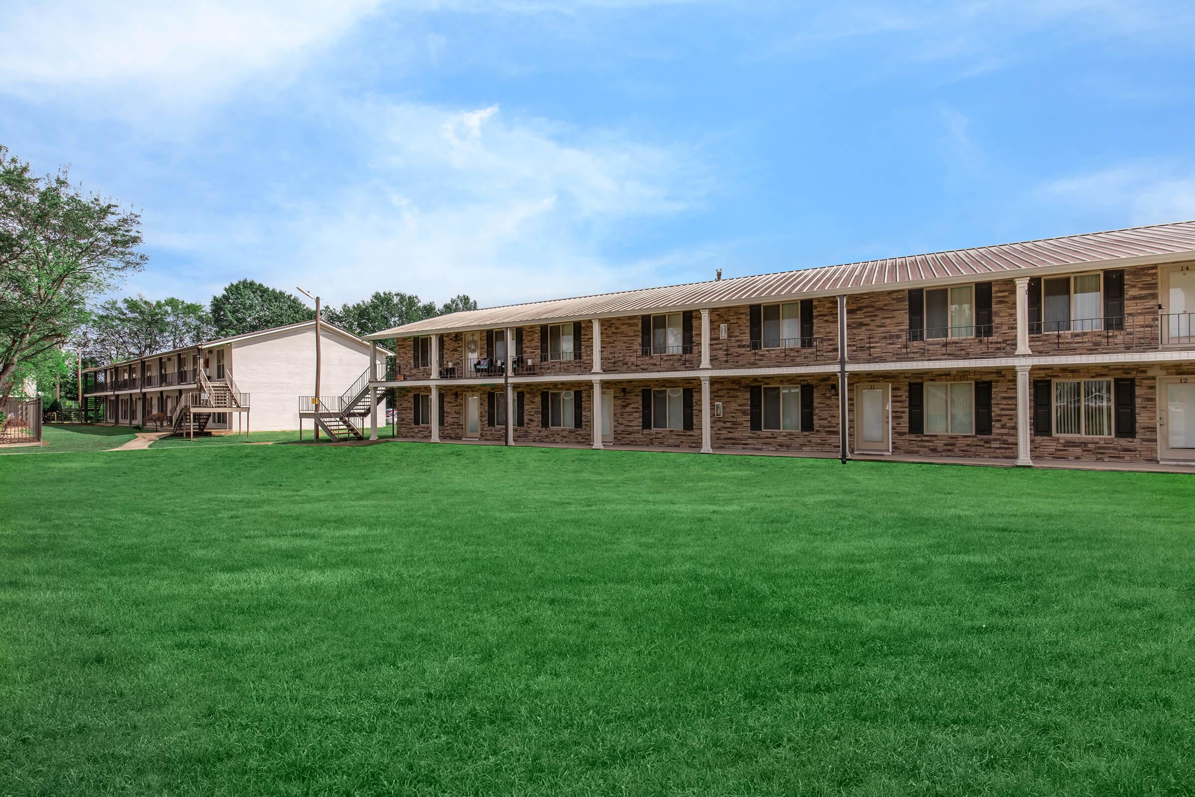 A view of a two-story apartment complex with a landscaped green lawn in front. The building features a mix of brick and siding exteriors, with balconies and multiple entrances. The sky is clear with some clouds, and there are trees in the background.