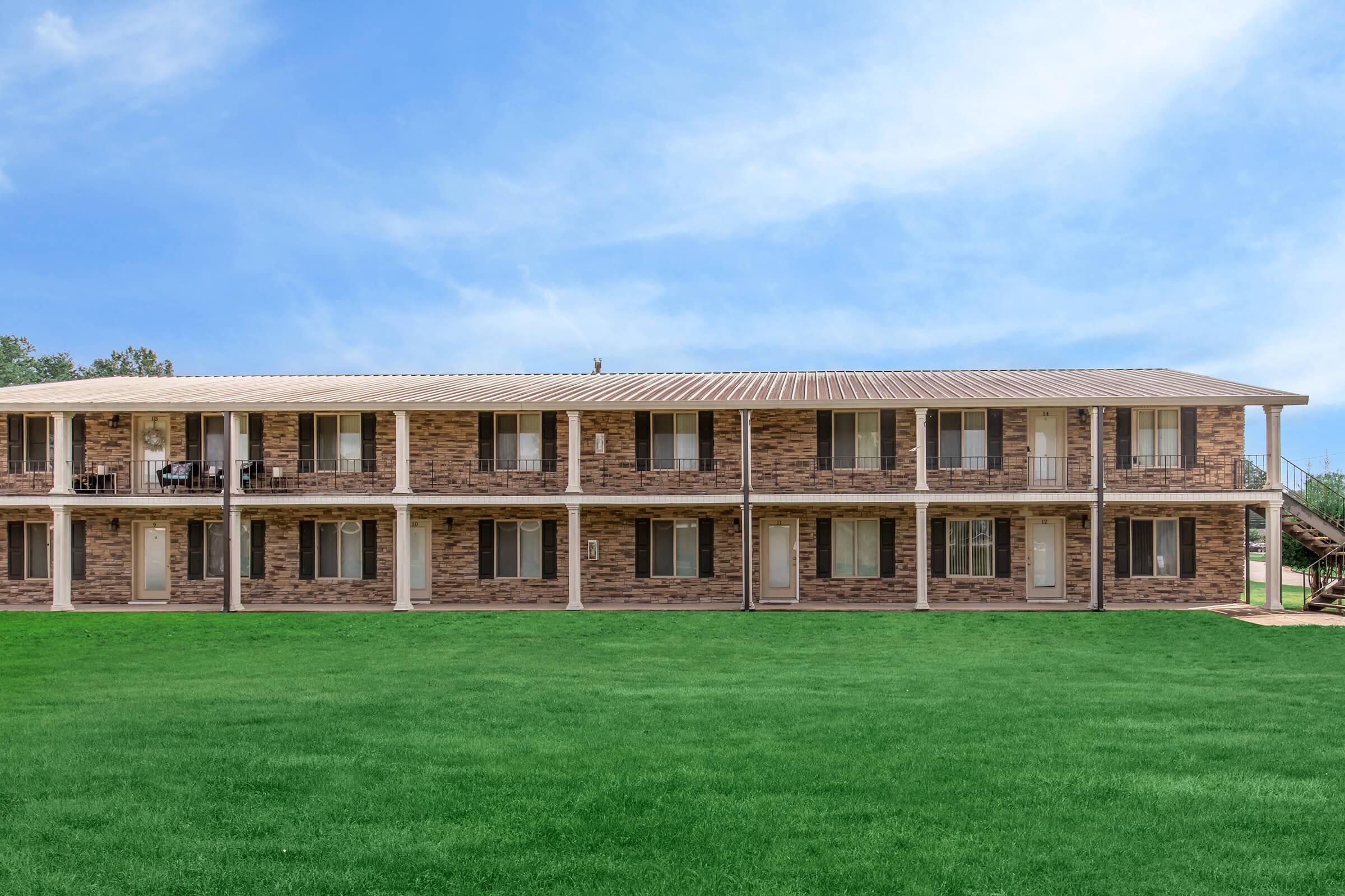 A two-story brick building with multiple outdoor balconies and windows. The structure is set against a clear blue sky and a green lawn in the foreground, creating a neat and inviting appearance.
