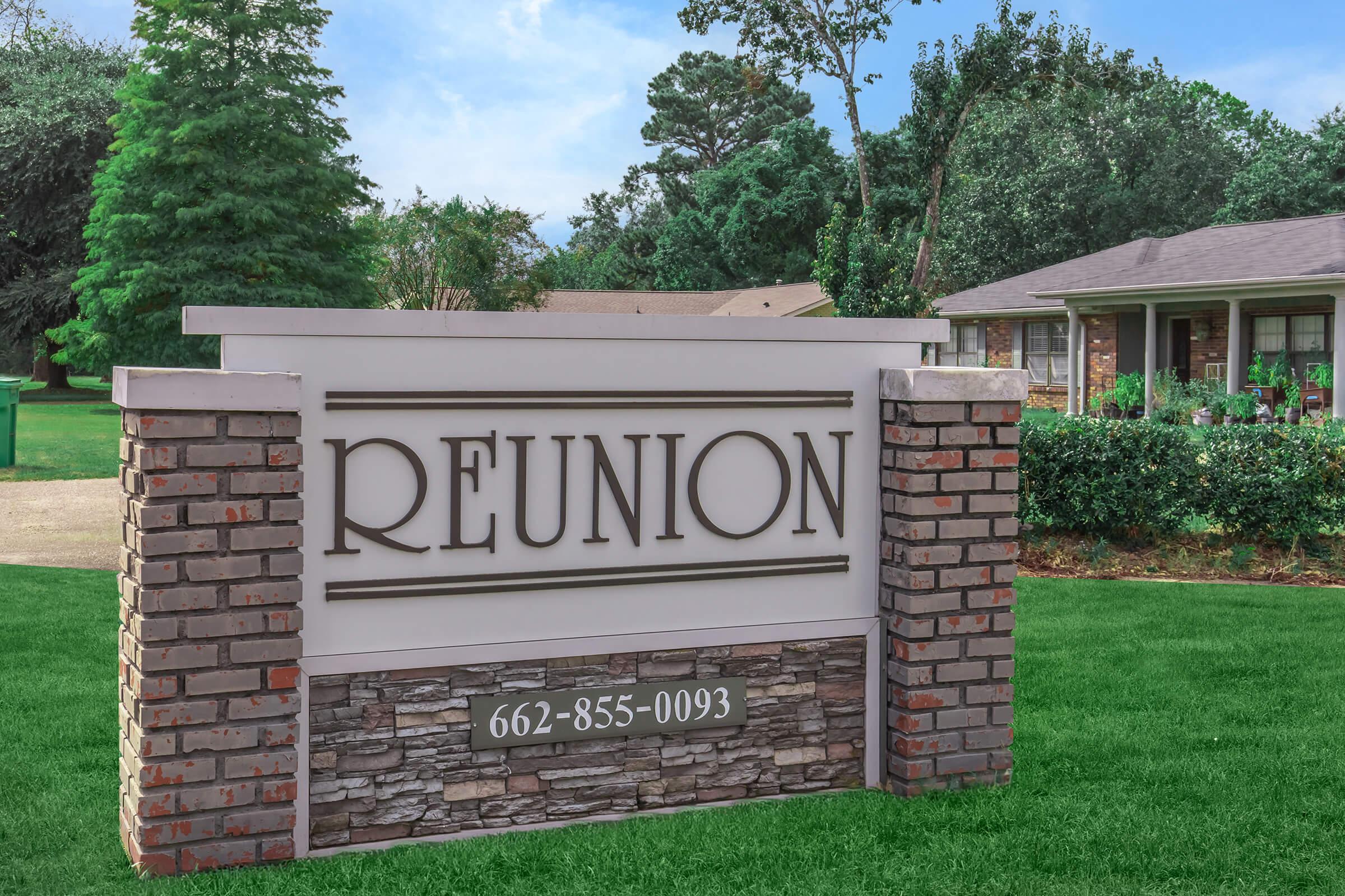 A sign labeled "REUNION" with a phone number, set against a backdrop of a green lawn and residential homes. The sign features a combination of brick and stone elements, and is surrounded by trees and shrubs, indicating a welcoming community setting.