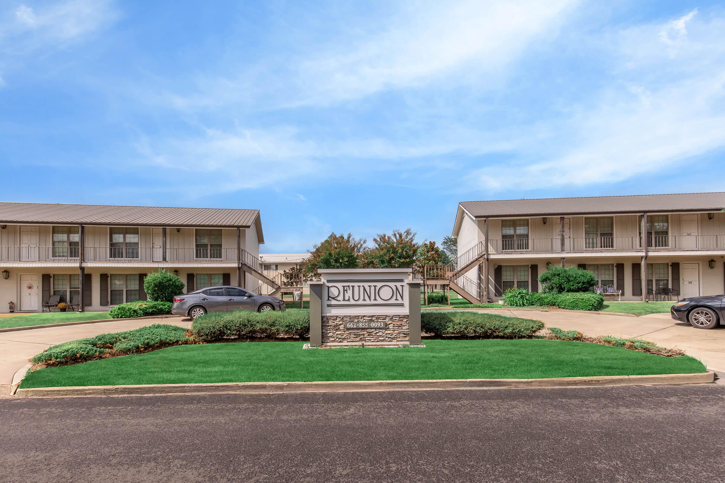 A view of the REUNION apartment complex featuring two-story buildings with balconies, well-maintained landscaping, and parking areas. The entrance sign is prominently displayed, surrounded by green grass and shrubs under a clear blue sky.