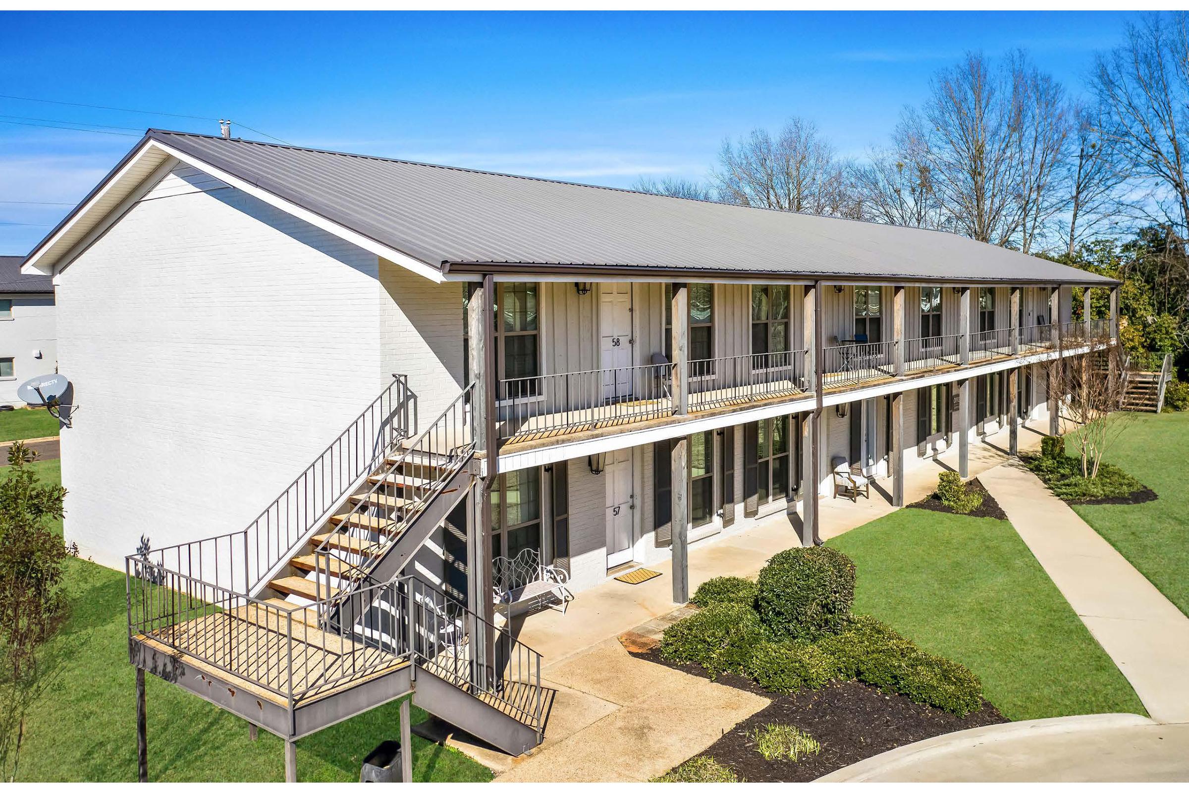 A two-story white motel with a metal roof, featuring multiple balcony suites. Stairs on the left lead to the second floor, with green grass and trees surrounding the building. Clear blue sky enhances the bright and welcoming appearance of the motel.