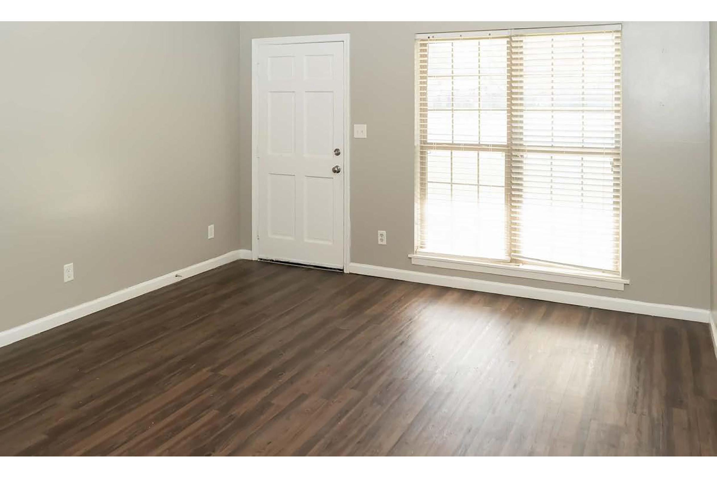 A well-lit, empty room featuring gray walls and a wooden floor. A window with horizontal blinds allows natural light to enter, while a white door is visible to the left. The space appears clean and ready for furniture placement.