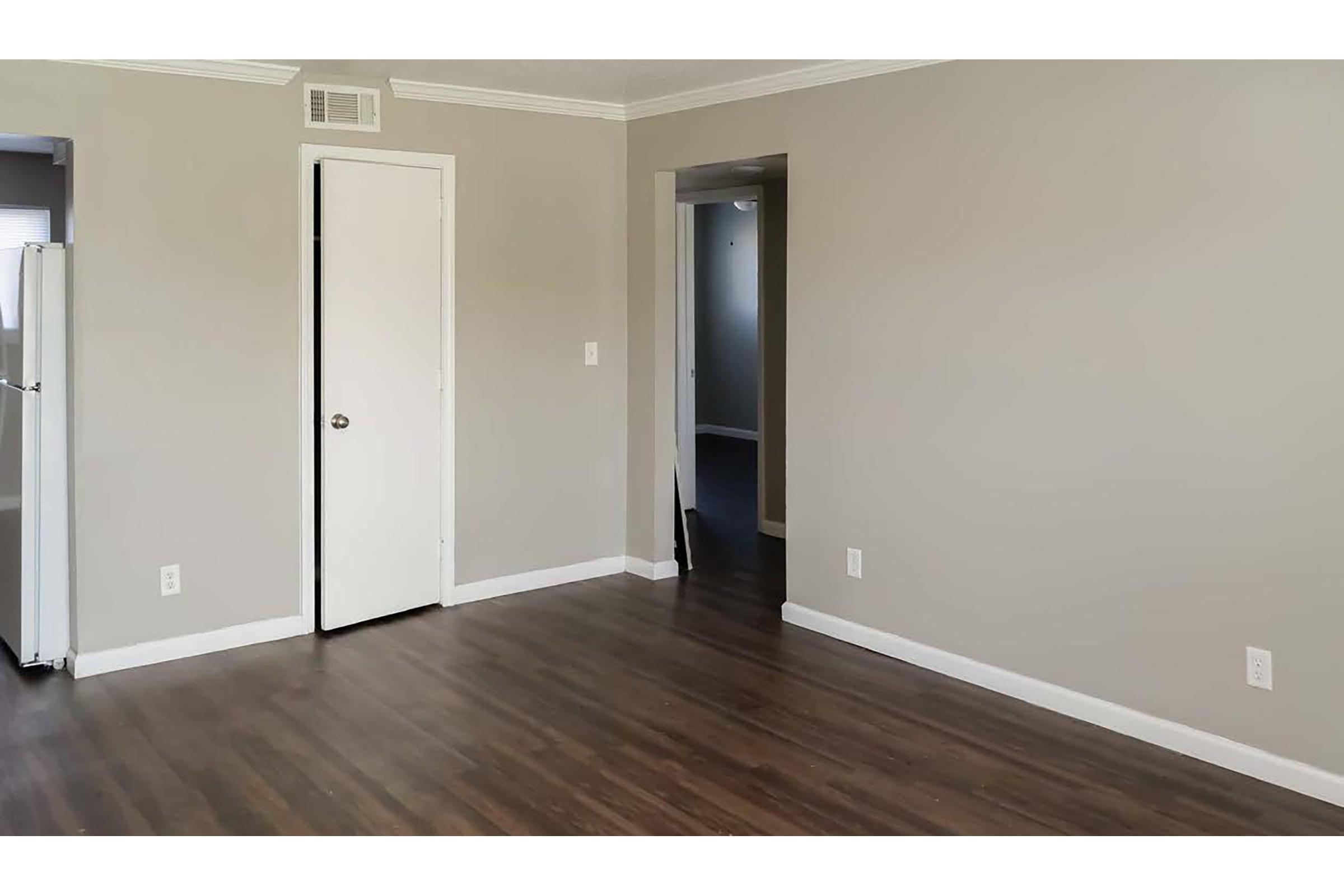 Empty room with light gray walls and dark wood flooring. A closed white door on the left leads to a closet or bathroom. A doorway on the right opens to another room, creating a sense of openness. Natural light brightens the space, enhancing its contemporary feel.