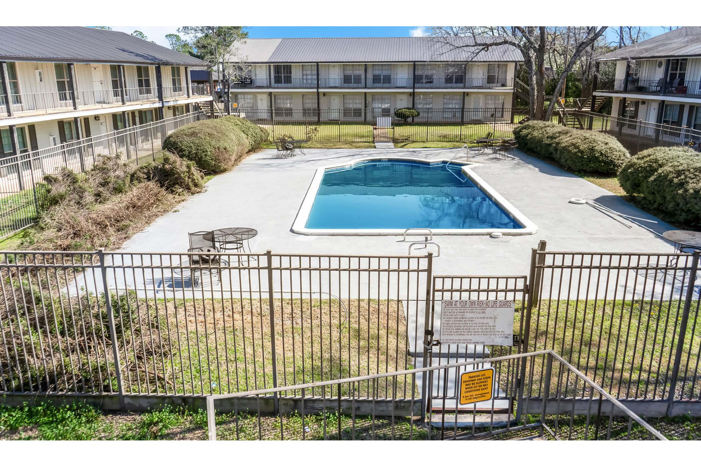 An outdoor swimming pool surrounded by a fenced area, with lounge chairs and tables nearby. The pool is clean and reflecting the blue sky. Surrounding the pool are neatly trimmed bushes, and apartment buildings are visible in the background, indicating a residential complex. A sign is posted near the entrance to the pool area.