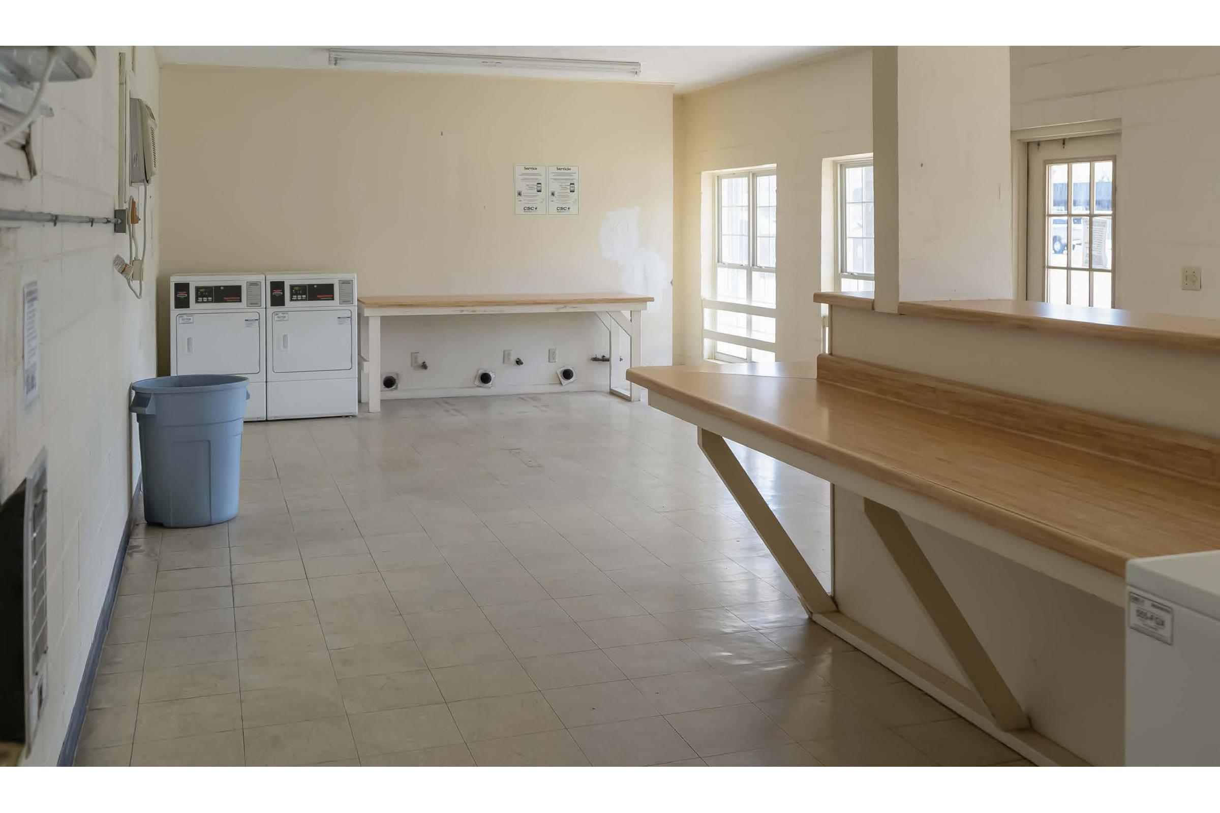 A spacious, well-lit laundry room featuring two washing machines and a dryer against one wall, countertop space for folding laundry, and a blue trash can. Large windows provide natural light, and the floor is tiled with a neutral color. Overall, the space appears clean and organized.