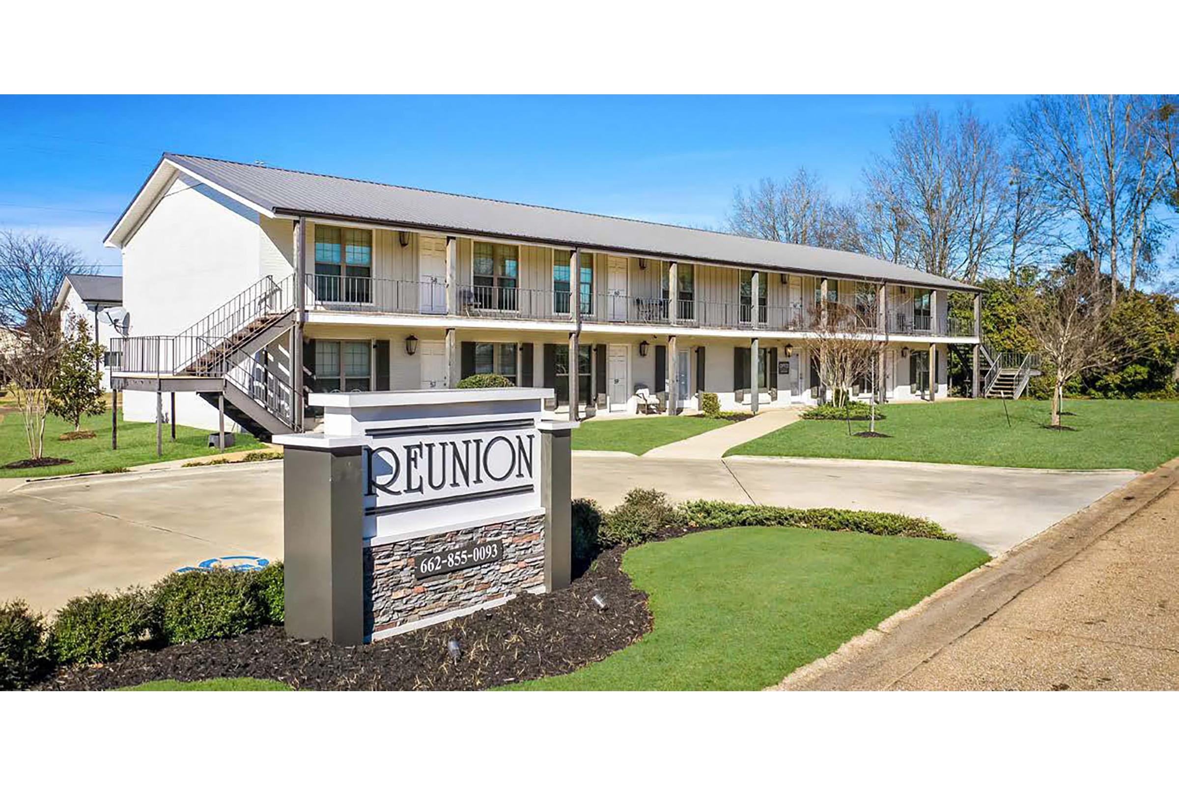 Two-story building with a modern design, featuring a sign that reads "REUNION" prominently in the foreground. The property is surrounded by well-maintained grass and landscaping. A clear blue sky is visible above, indicating a sunny day.