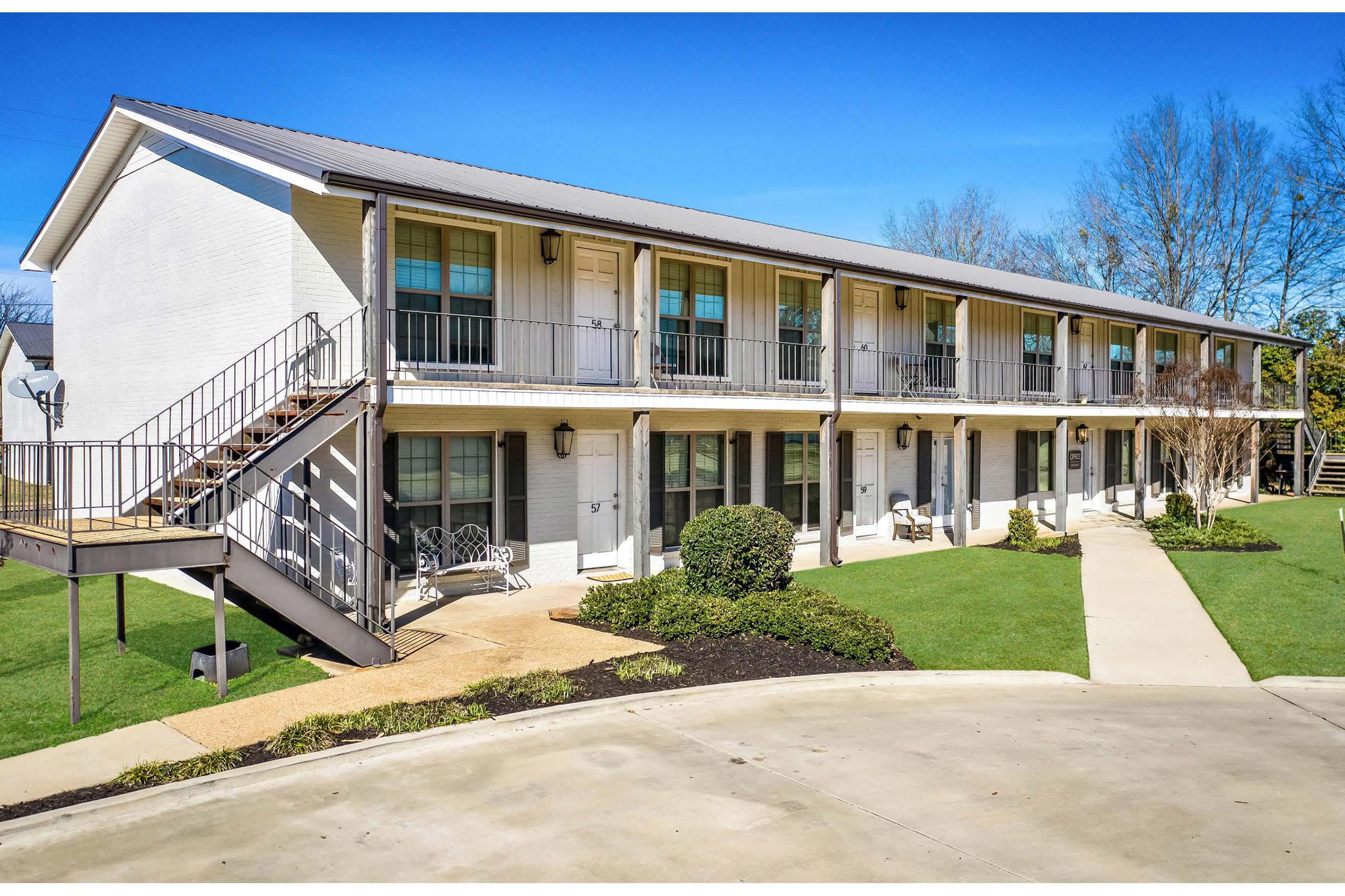 A two-story white building with balconies, featuring a staircase on the left side. Lush green grass and shrubs surround the path leading to the entrance. Clear blue sky above adds to the bright atmosphere of the scene.