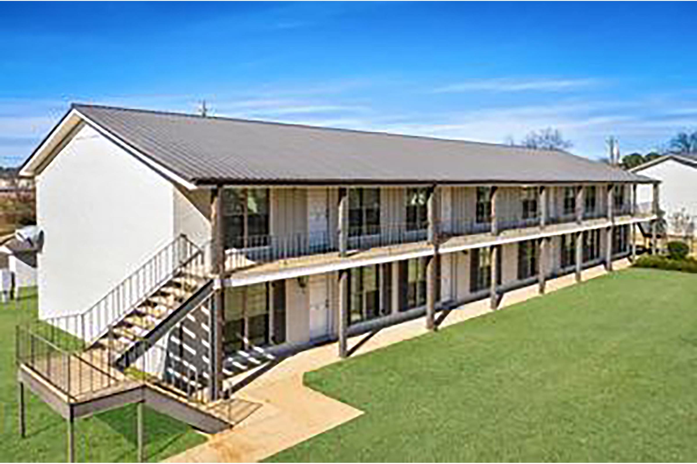 A two-story building with a metal roof, featuring multiple balconies. The structure has a grassy area in front and outdoor stairs leading to the upper level. Clear blue sky and well-maintained surroundings are visible.