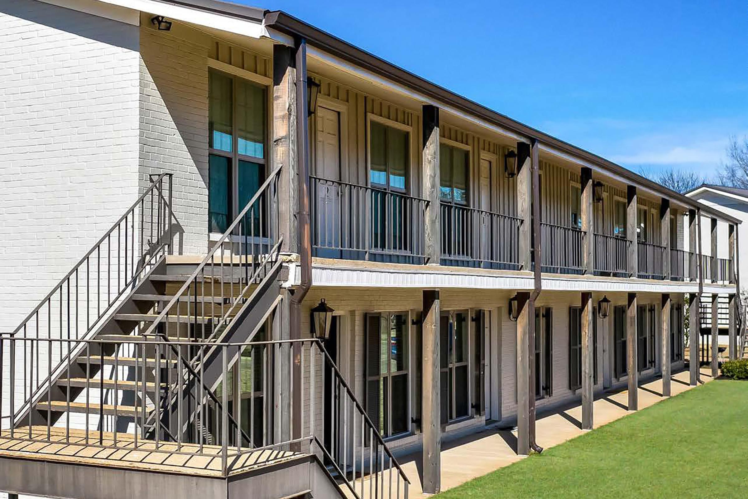 A two-story building with a modern design featuring several balconies and large windows. The exterior is painted white, and there's a staircase leading to the upper level. The surrounding area is green grass under a clear blue sky.