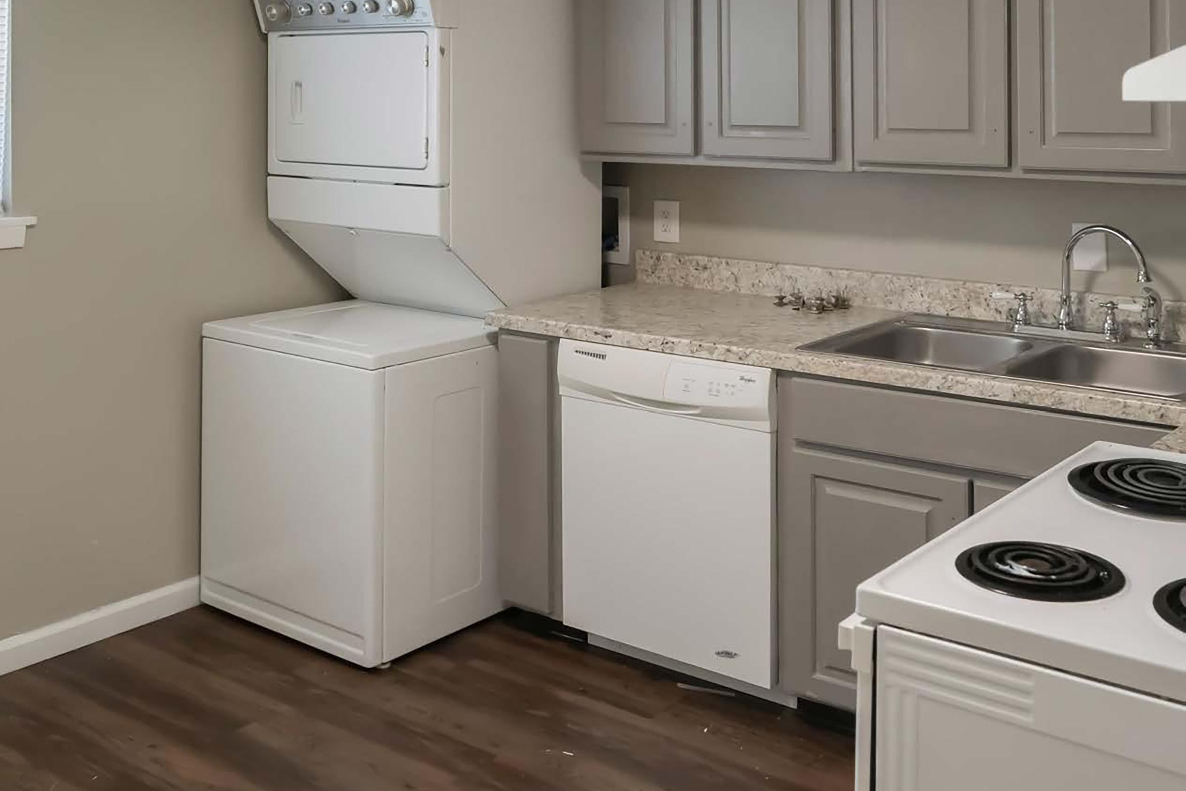 A modern kitchen featuring a stacked washer and dryer unit on the left, a dishwasher next to it, a double sink with a chrome faucet, and a stove with two burners. The cabinetry is light gray, and the countertop is a speckled design. The flooring is a dark wood laminate.