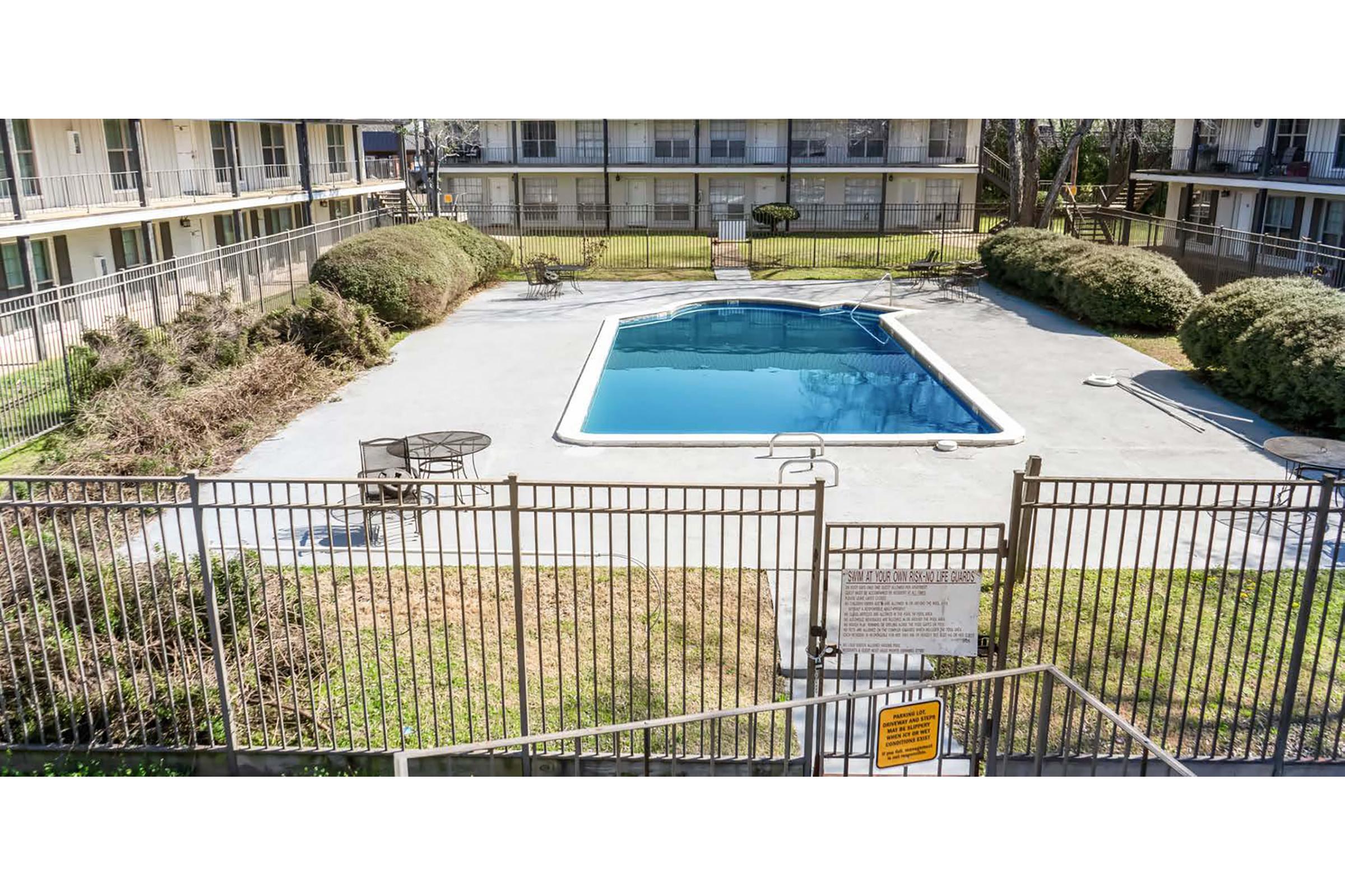A fenced swimming pool area at a hotel or apartment complex, featuring a clear blue pool surrounded by concrete and grass. There are chairs and a table nearby. The pool area is bordered by a low wall and a gate, and there are shrubs in the background, indicating a well-maintained outdoor space.