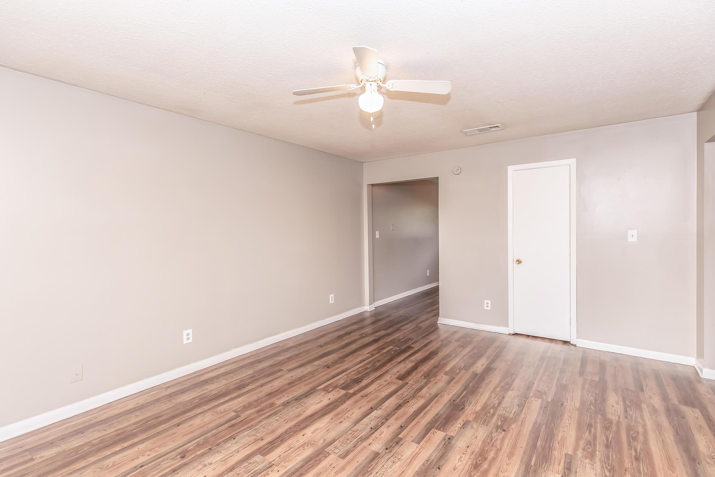A spacious, empty living room with laminate flooring and a ceiling fan. The walls are painted in a light neutral color, and there is a closed white door on the right. A doorway leads to another room in the background, creating an open and airy feel.