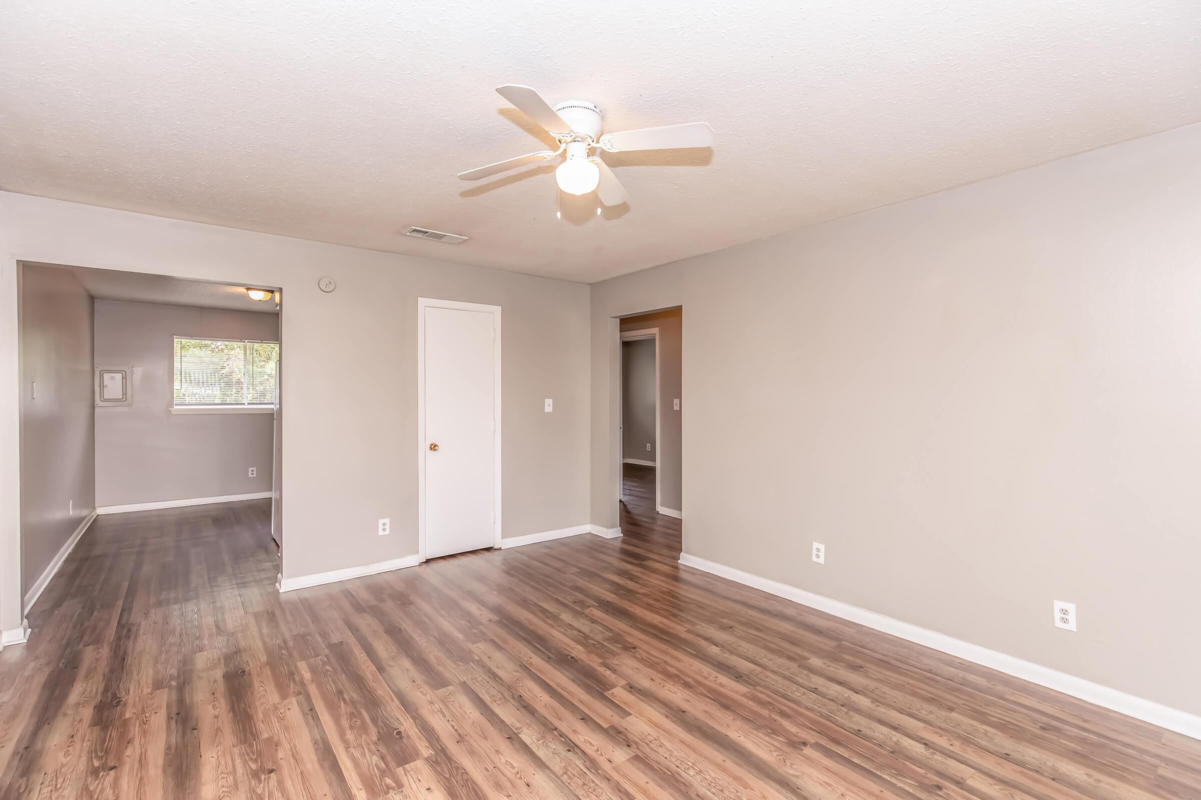 A spacious, light gray living room with a ceiling fan and laminate wood flooring. The room features a closed door and an open doorway leading to another room. Natural light enters through a window, creating a welcoming atmosphere. The walls are neutral, enhancing the sense of openness.