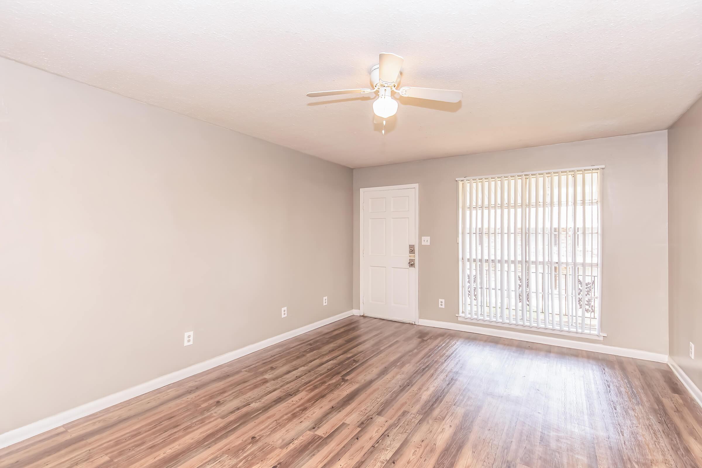 A spacious, empty living room featuring light-colored walls, hardwood flooring, and a ceiling fan. A door is visible on the left side, and a window with vertical blinds allows natural light to enter. The room is ready for furnishing and personalization.