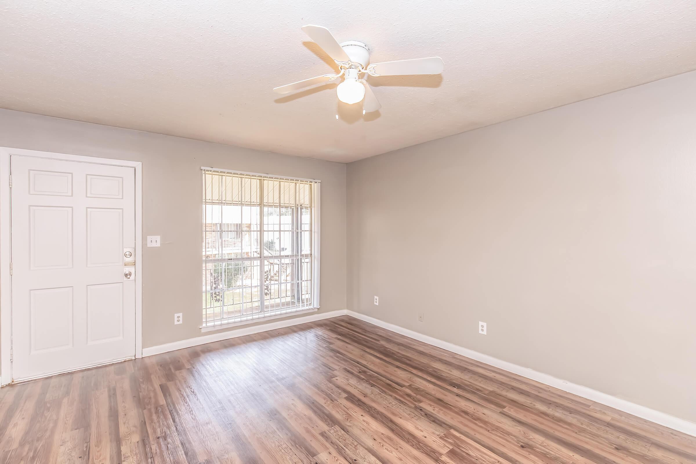 A spacious room with light-colored walls and a ceiling fan. A large window lets in natural light, and the floor features wood-like laminate. The entrance door is visible on the left, and the overall atmosphere is bright and airy, suitable for living or office use.