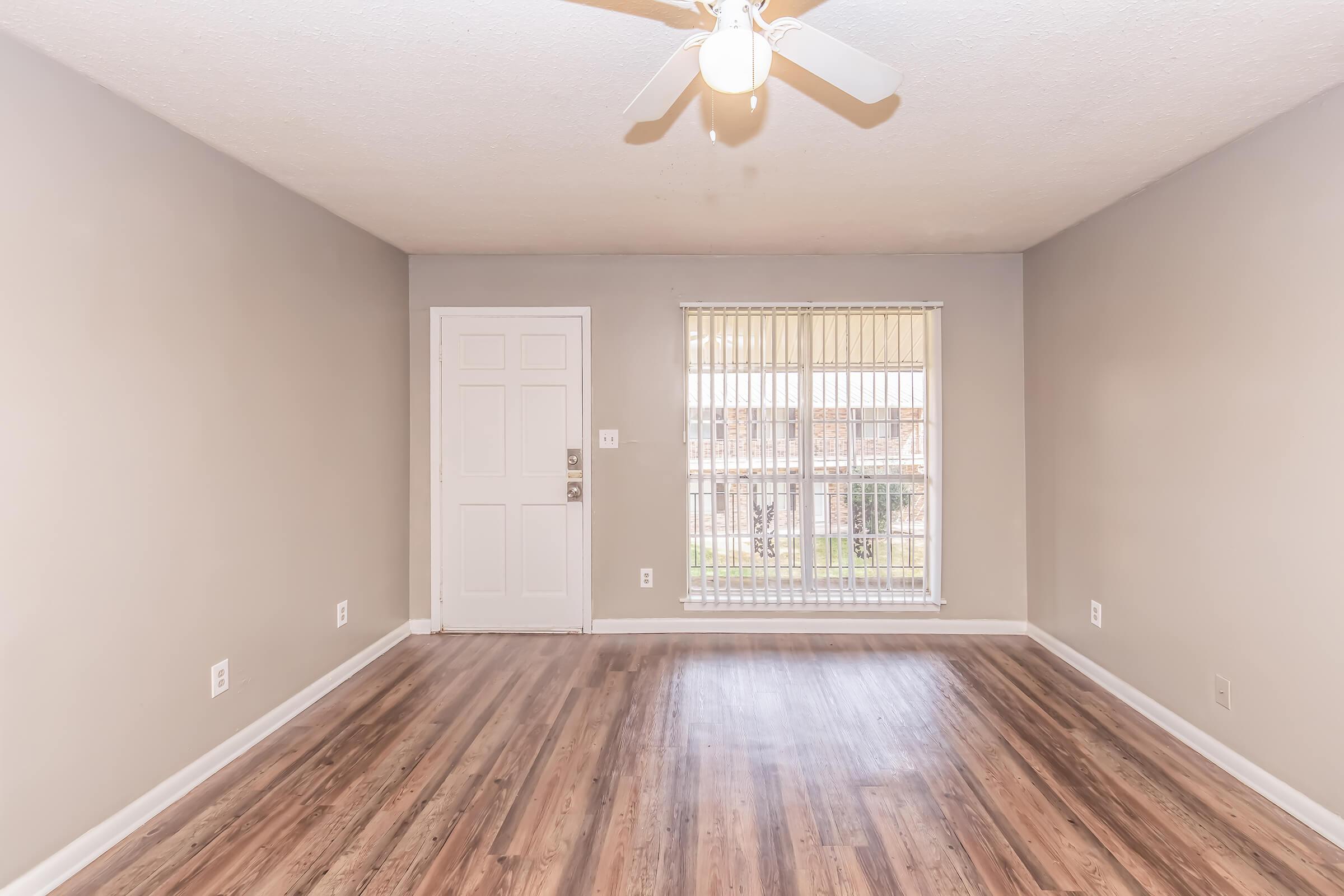 A spacious, empty living room featuring light-colored walls, hardwood flooring, a ceiling fan, and a large window with blinds. The room has a front door visible on the left side, allowing natural light to fill the space.