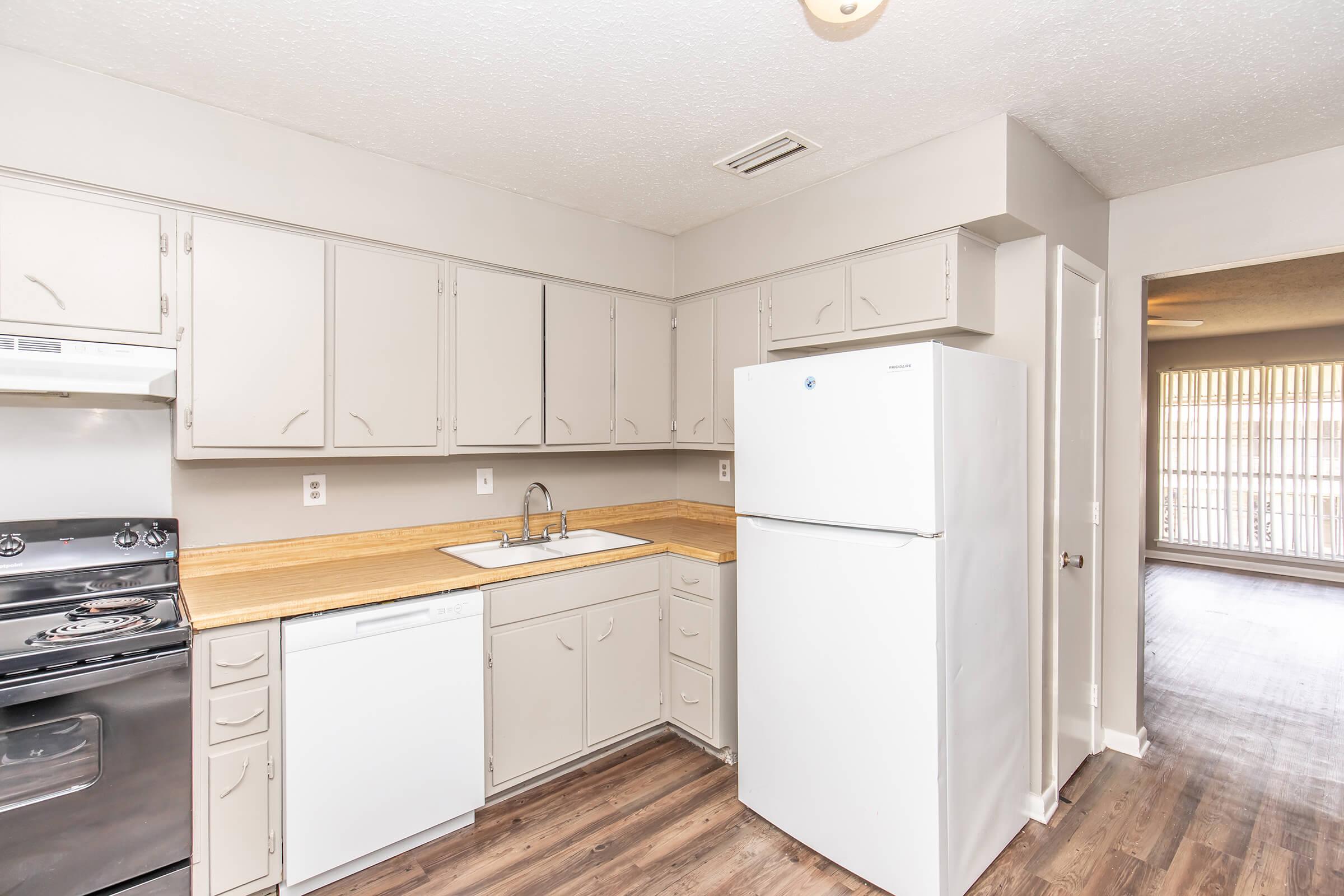 A kitchen with light gray cabinets, a white refrigerator, a stainless steel stove, and a sink. The countertop is made of wood. The flooring is a dark wood laminate. In the background, there is a view of a living space with large windows and shades, providing natural light.