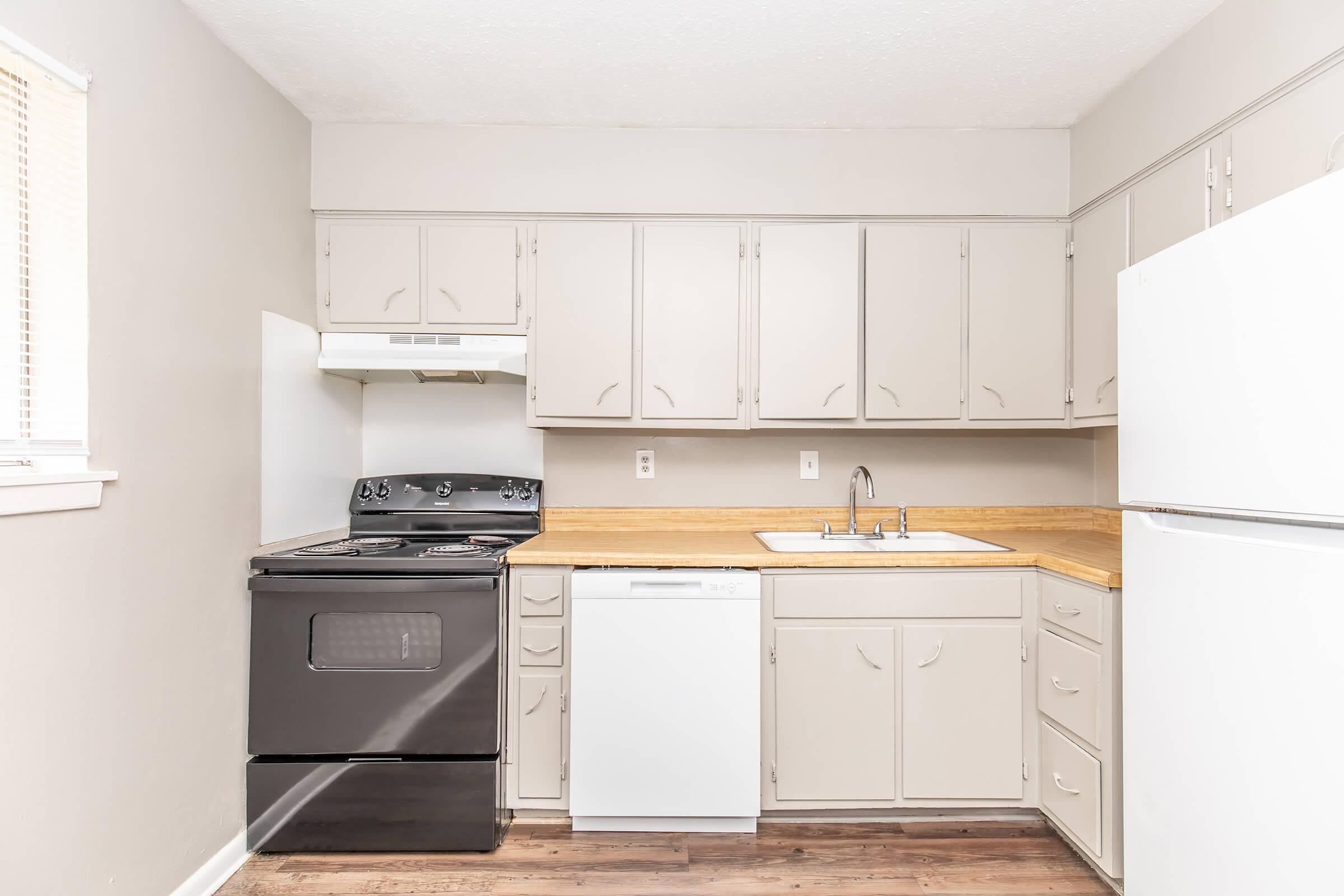 A clean and modern kitchen featuring light gray cabinets, a black stove, a white dishwasher, and a double sink. The countertop is wooden, and the walls are painted in a neutral tone. Natural light comes in through a window, enhancing the overall spacious feel of the room.