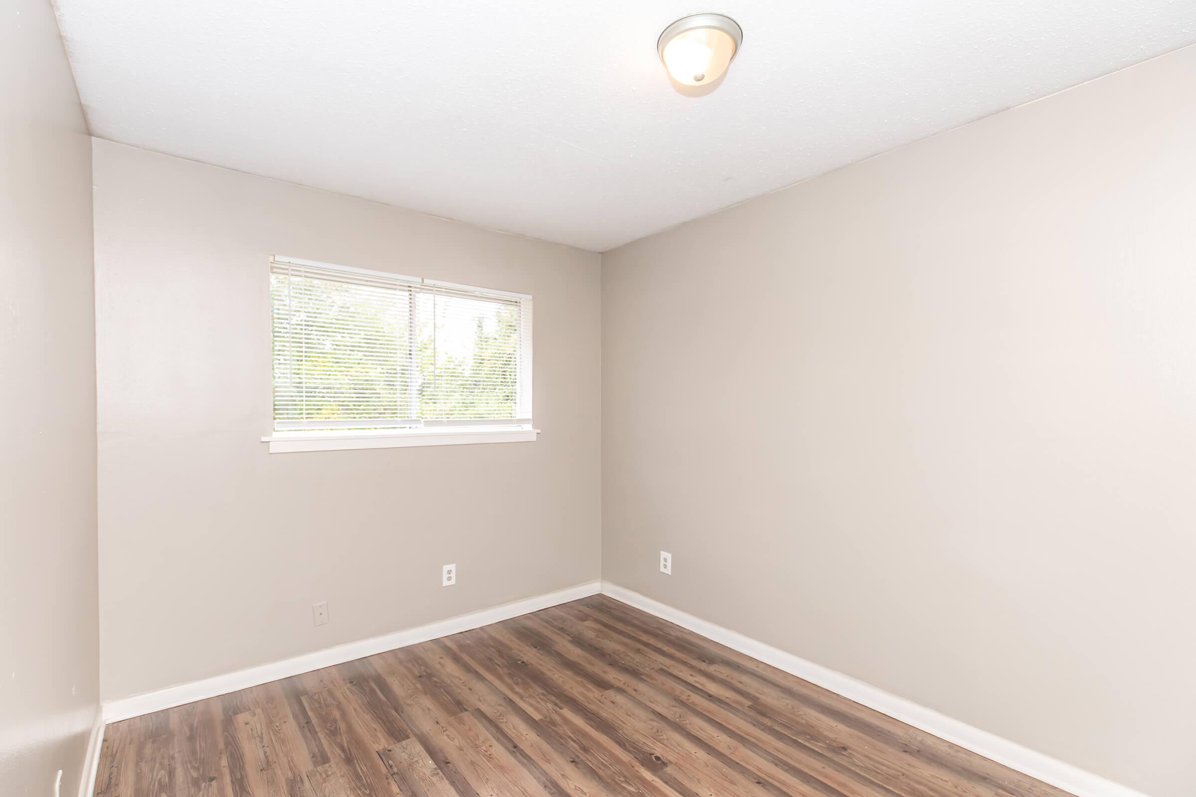 A small, empty room with light brown walls, a single window with blinds allowing natural light, and a ceiling light fixture. The floor is covered with light wooden laminate. No furniture or decorations are present, creating a minimalist appearance.