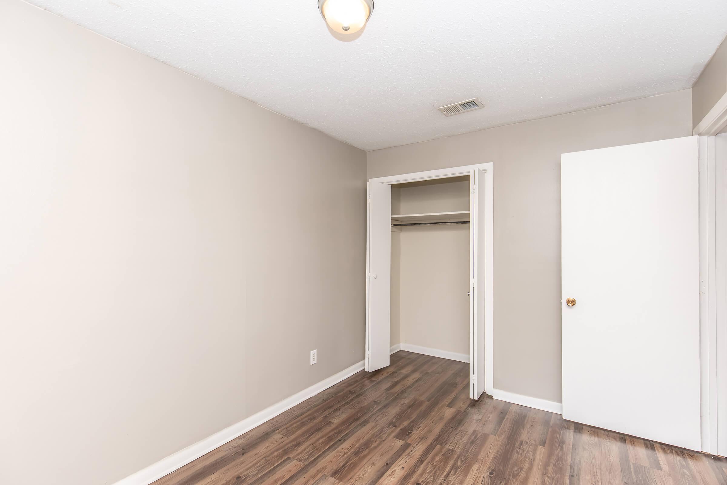 Empty room with beige walls and hardwood flooring. A closet is visible with an open door, and there is a light fixture on the ceiling. The space appears bright and uncluttered, offering a simple and neat environment.