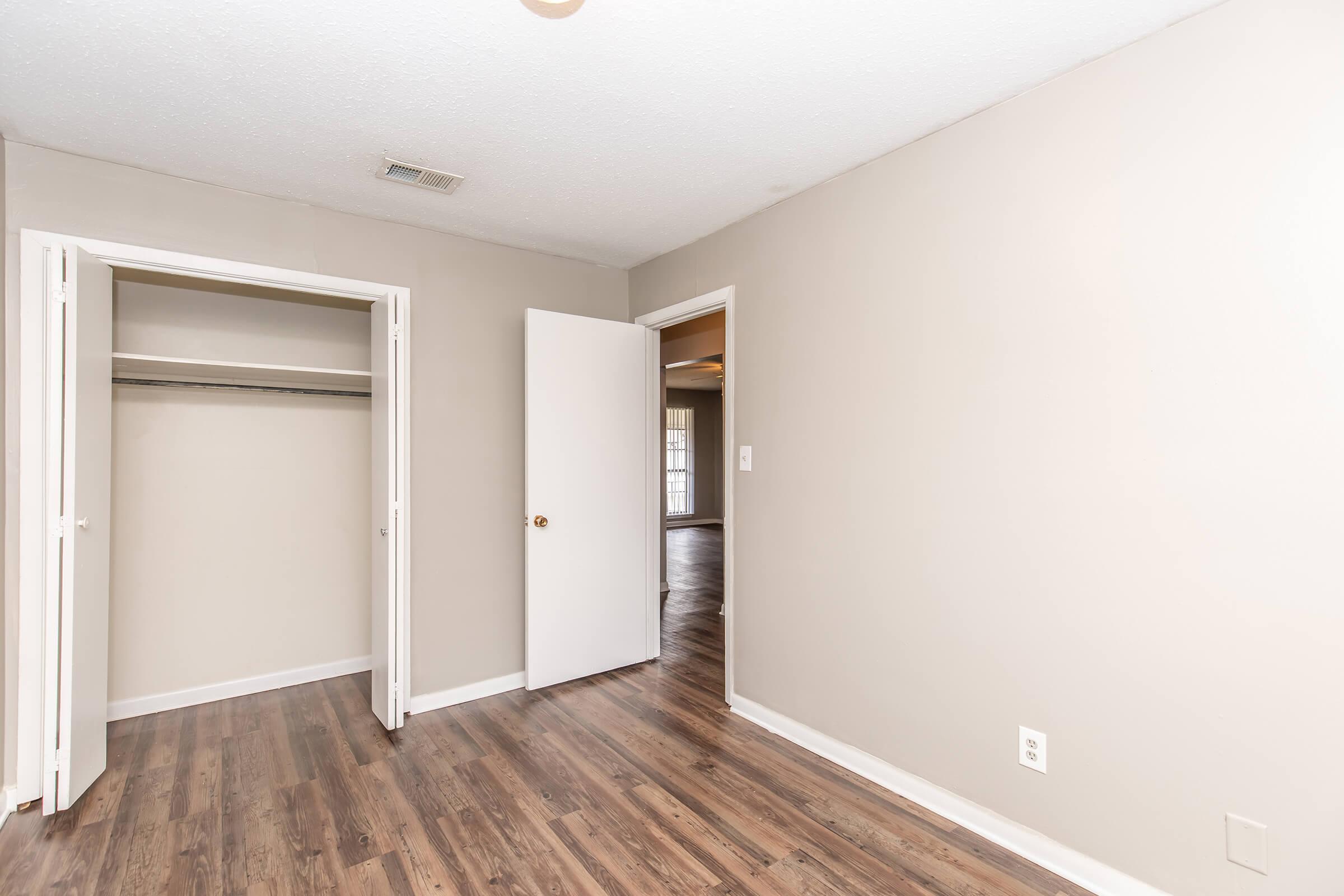 A room featuring light beige walls, a door leading to another area, and a closet with no doors. The floor is wood-like laminate, and the space appears clean and well-lit, suitable for various uses.