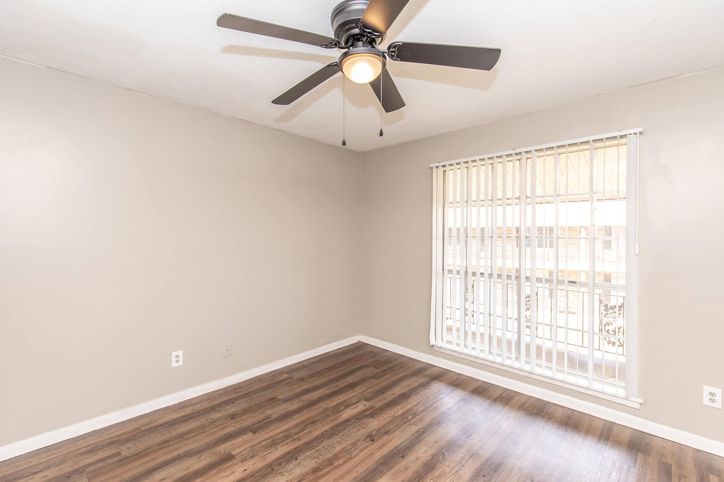 Empty room with light gray walls and a ceiling fan. There is a window with vertical blinds letting in natural light, and the floor is covered with laminate wood flooring. The space appears clean and ready for furniture.