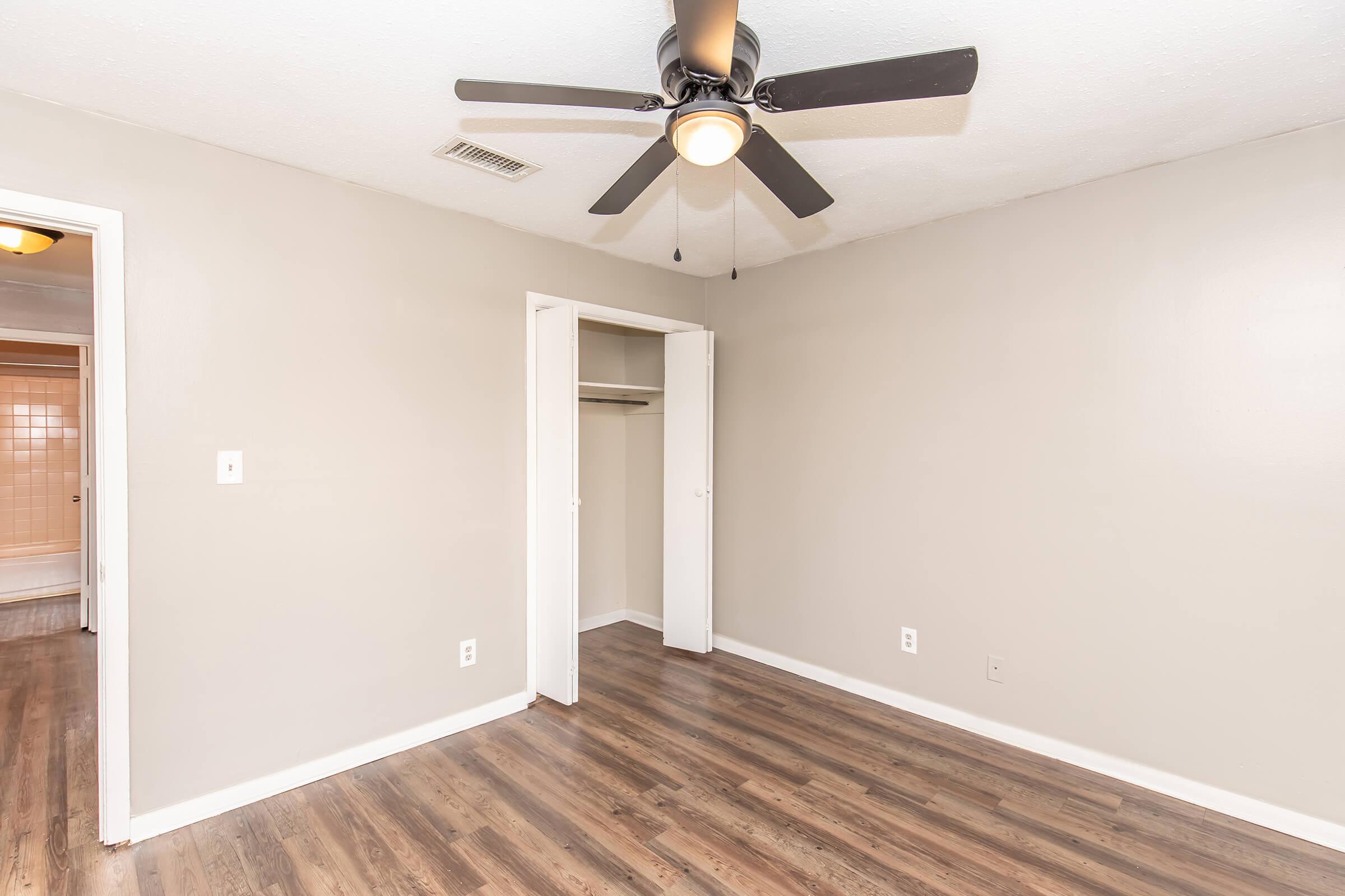 A clean, empty room featuring light gray walls and a ceiling fan. The room has a closet with double doors on one side, and polished wooden flooring throughout. A doorway leads to a bathroom visible in the background. The space is well-lit and appears spacious.