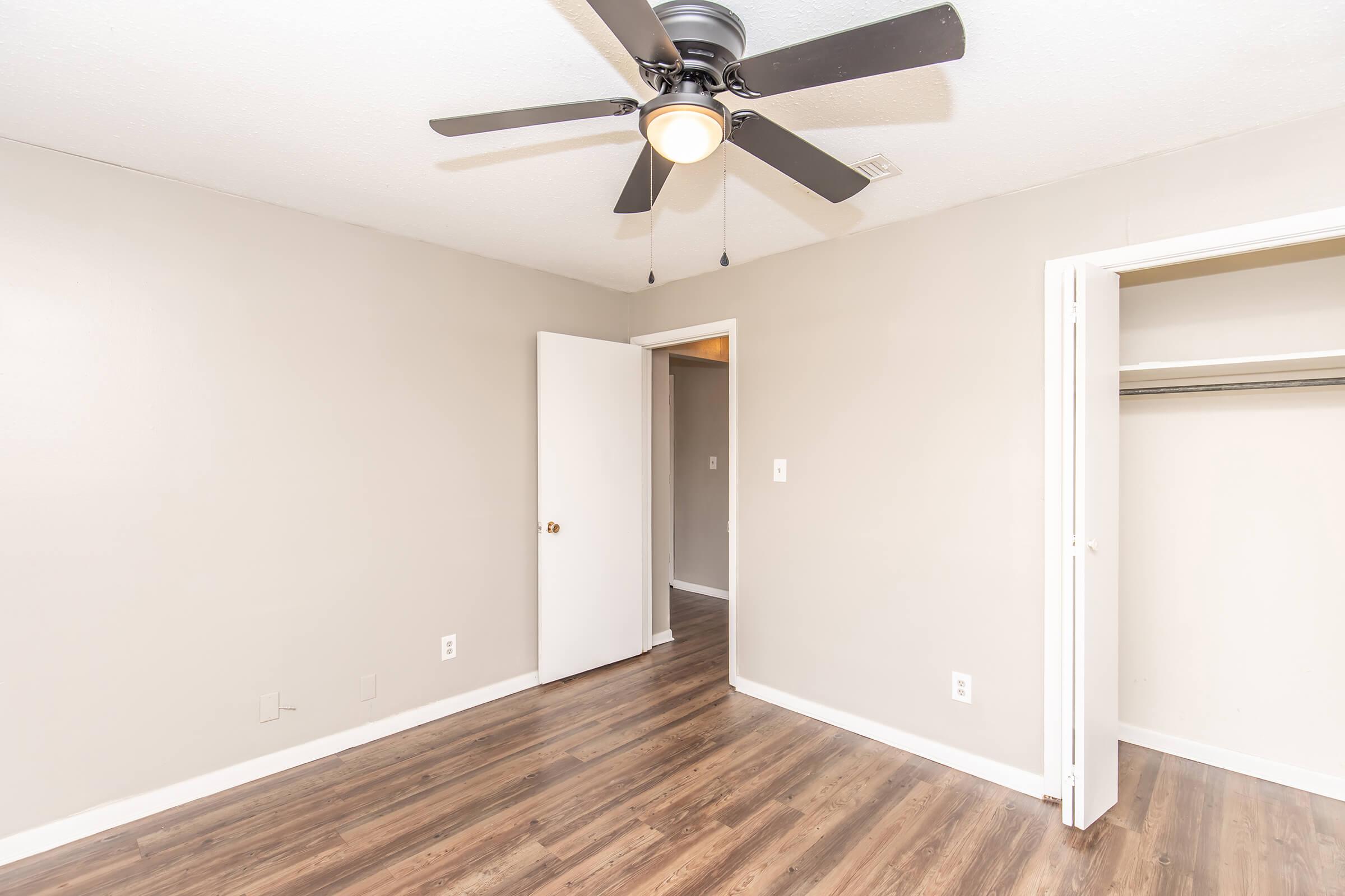 A simple room featuring a ceiling fan, beige walls, and hardwood flooring. There are two doors: one leading to a closet and another to a different space. The room is empty, creating a spacious and bright atmosphere.