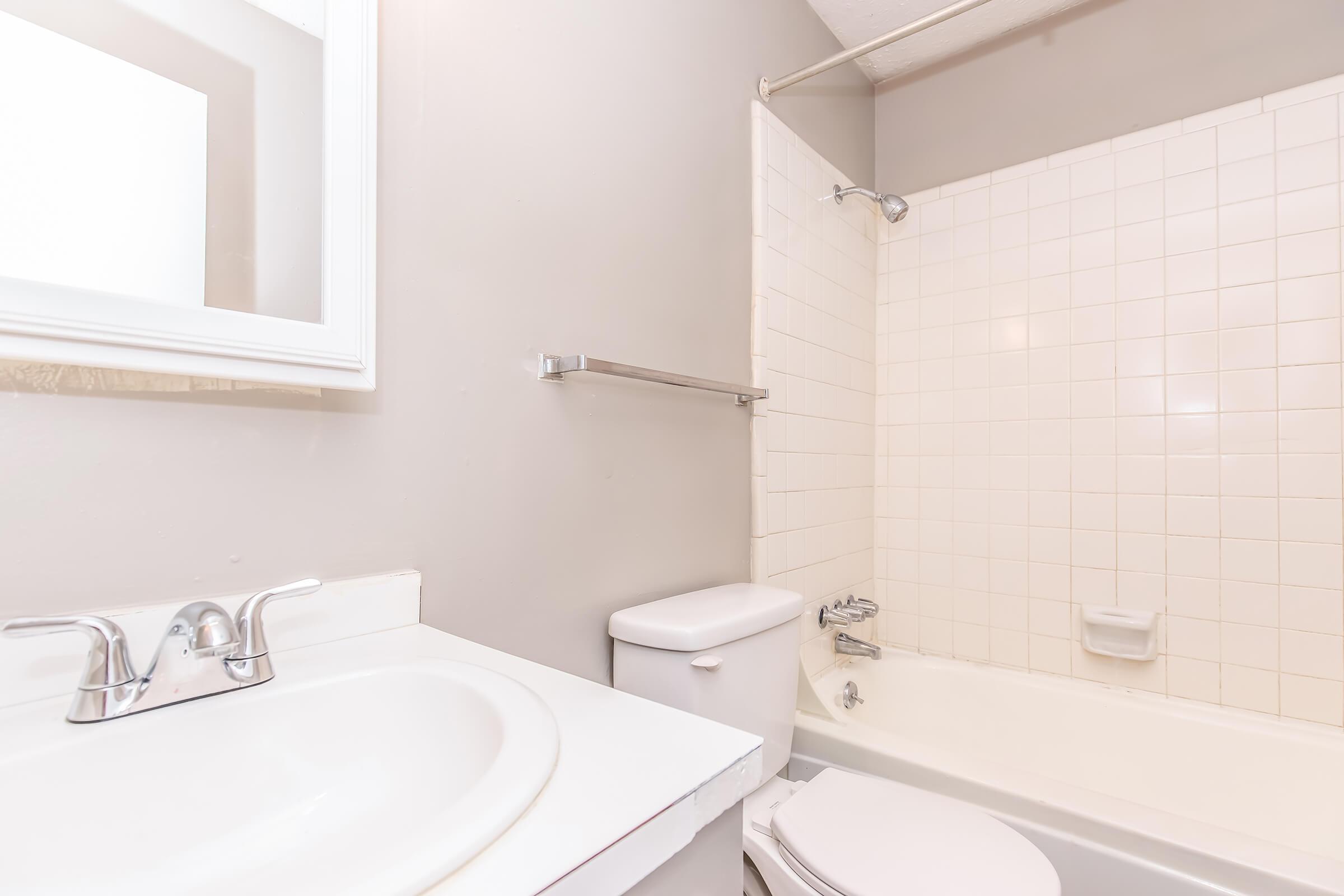 A simple bathroom featuring a bathtub with a shower, a white toilet, and a sink with a mirror above. The walls are painted light gray, and the tile is light-colored. The overall design is clean and minimalistic, with a focus on functionality.