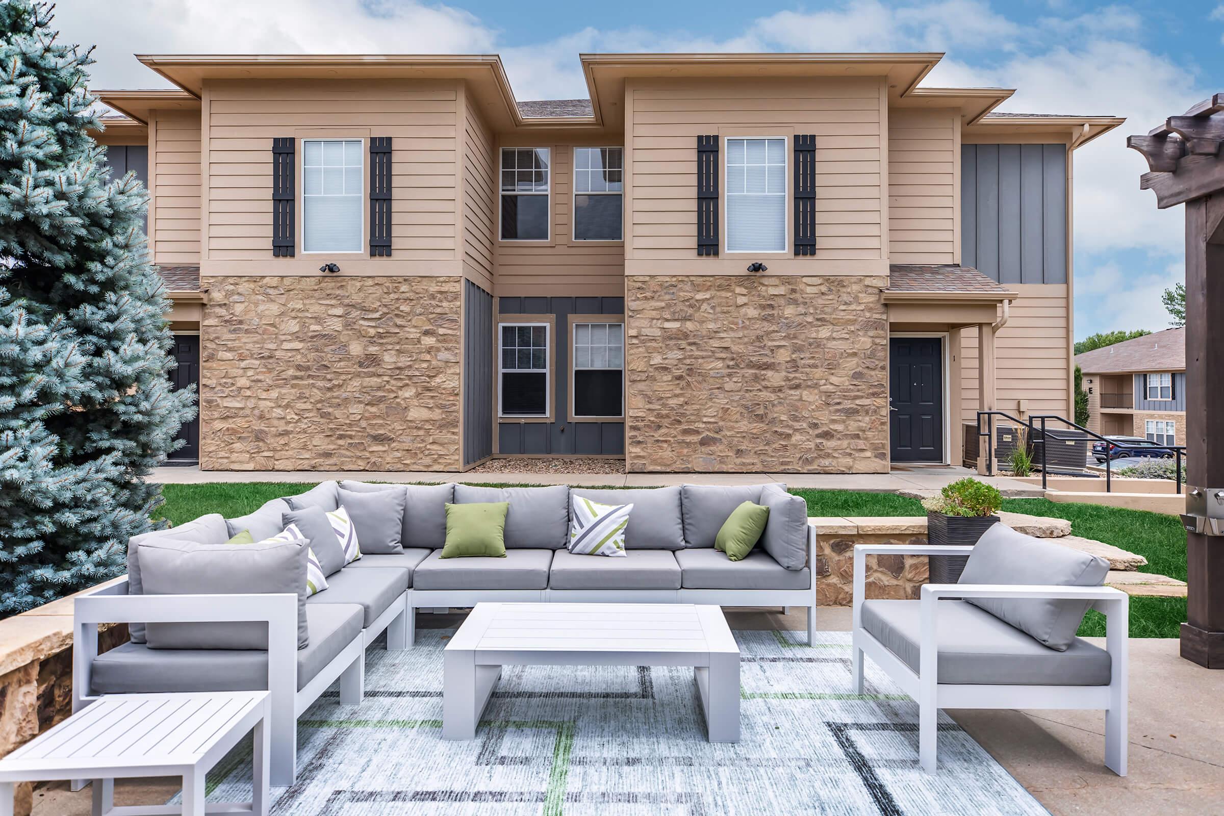 A modern outdoor seating area with a gray sectional sofa, accent pillows, and a white coffee table, set against a backdrop of a two-story building with stone and vinyl siding. Lush green grass and a light blue sky complete the scene.