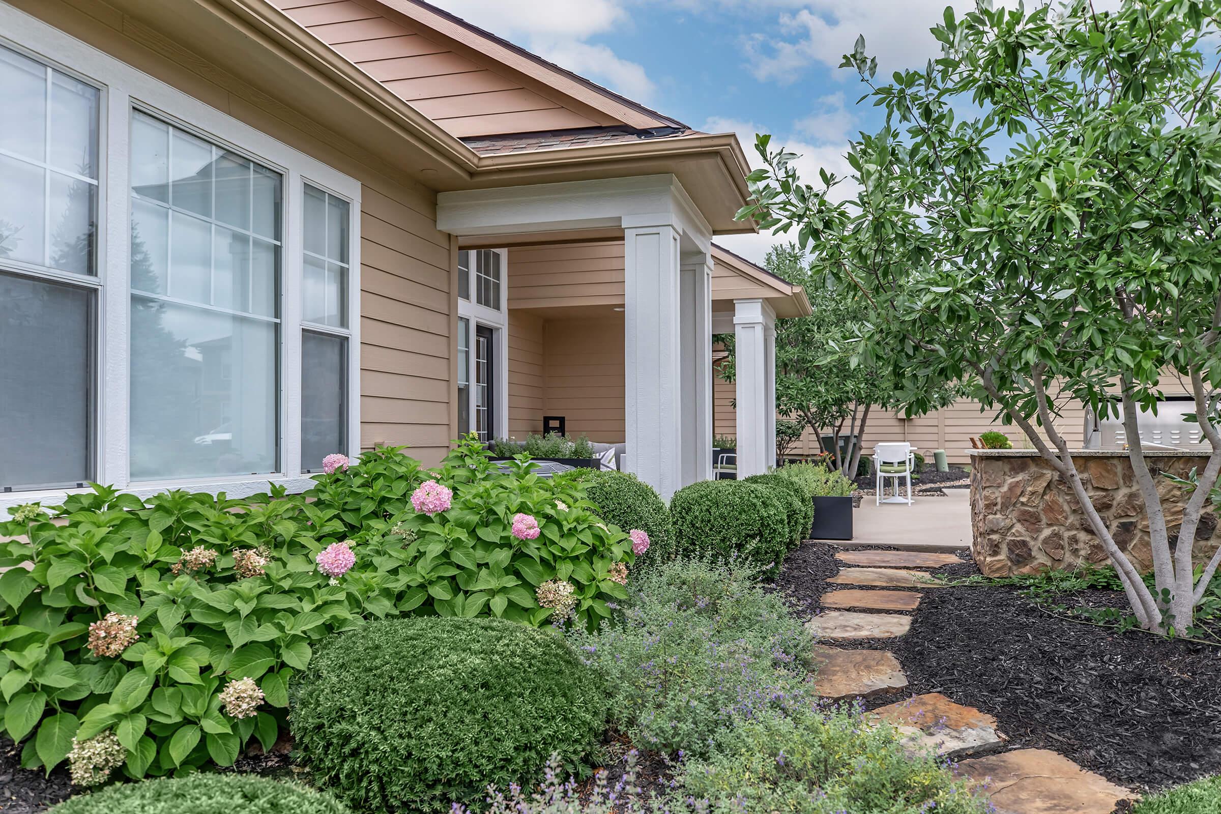 A well-maintained garden with lush green shrubs and blooming hydrangeas frames a residential home. The house features a light brown exterior and a covered porch with seating. A stone pathway leads through the vibrant landscaping, creating an inviting atmosphere in a serene outdoor setting.